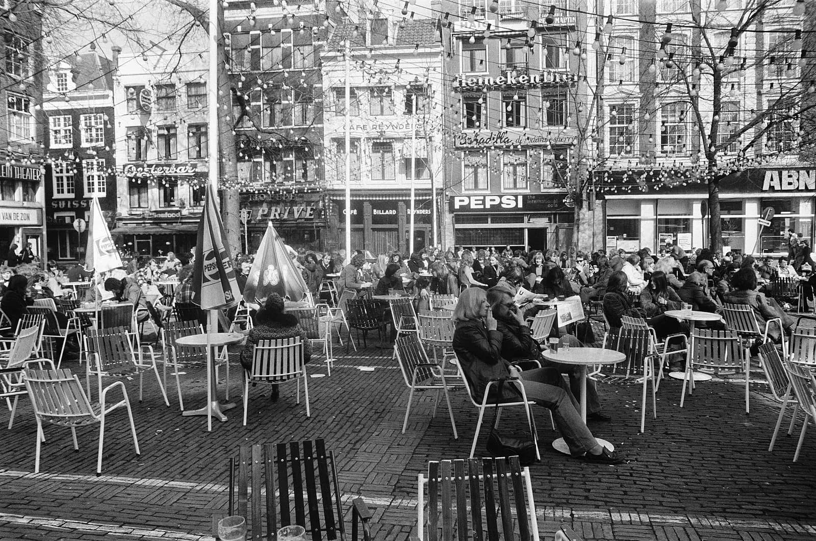 Black and white photograph of a crowded café terrace on a cobbled square, people in coats sitting on metal chairs at small round tables, with tall narrow buildings carrying signs for Heineken, Pepsi and a bank behind.