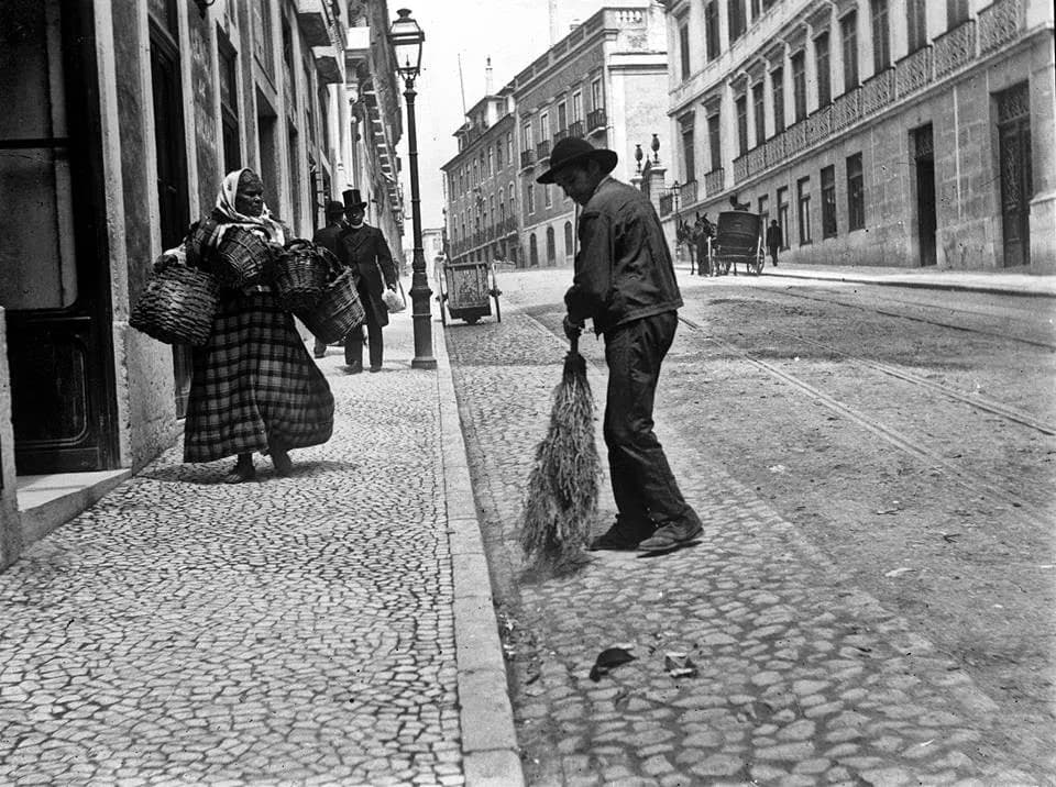 Black and white street scene: a woman in a long checked skirt and headscarf carries large baskets along a cobbled pavement while a man in a broad hat sweeps the street. A man in a top hat walks behind her. Tall stone buildings and a gas lamp line the street, and rails run along the road.