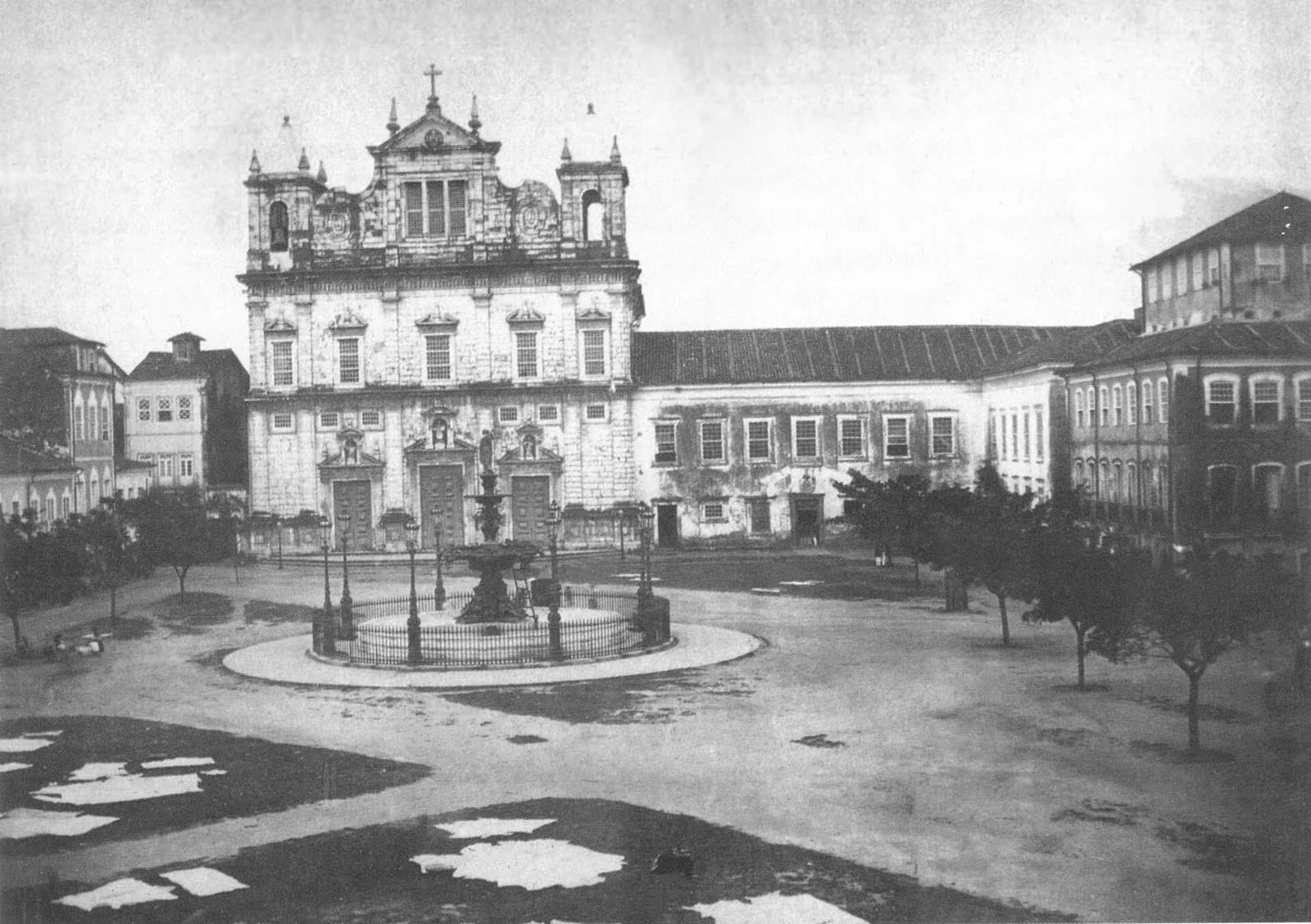 Old black and white photograph of a wide square with a fountain and young trees. Behind it stands a large white Baroque church with two short towers, joined to a long two-storey building.