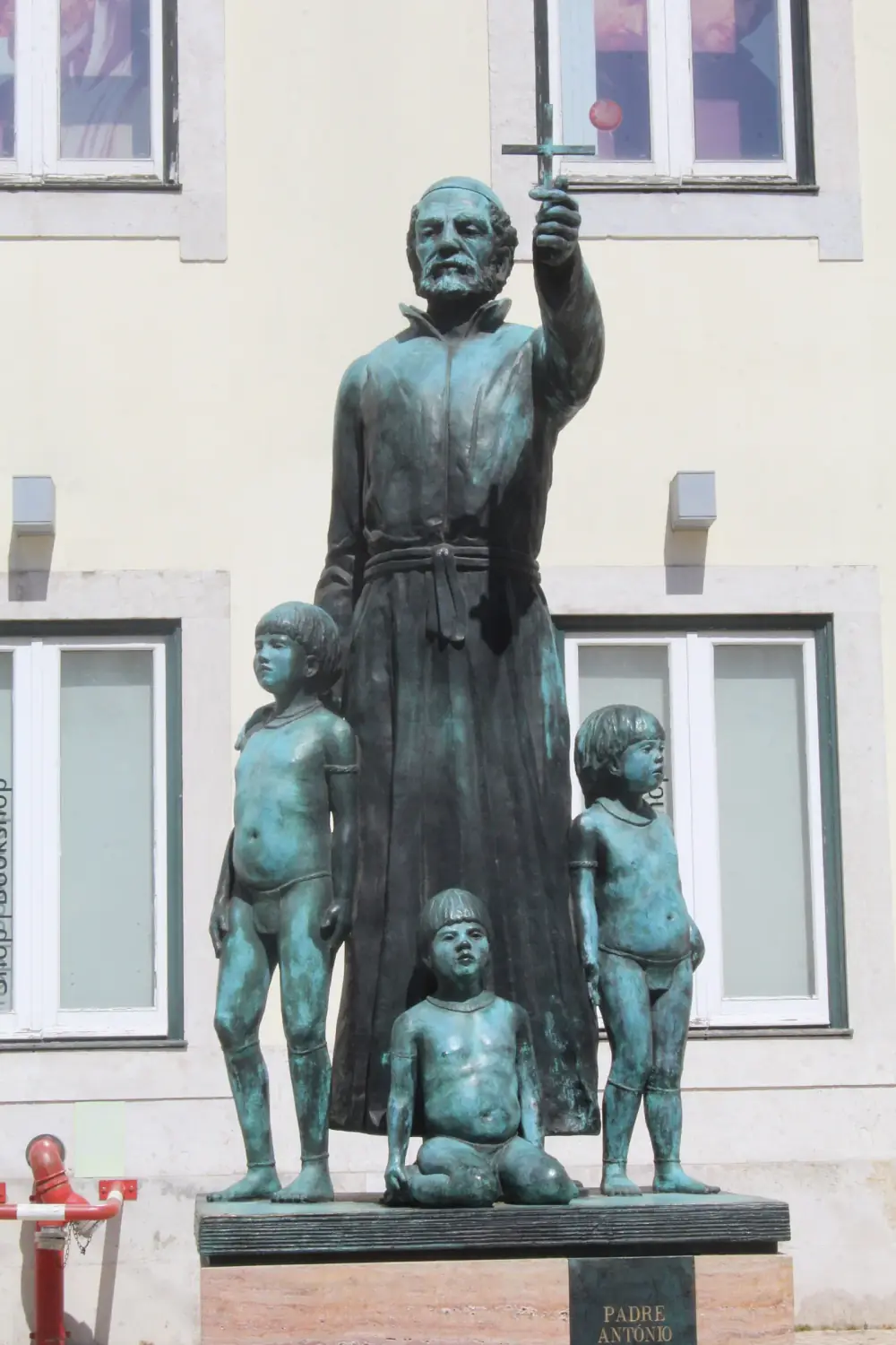 Bronze statue of a bearded priest in a long gown holding up a small cross, with three almost naked Indigenous children standing and kneeling at his feet, on a stone plinth in front of a pale building.