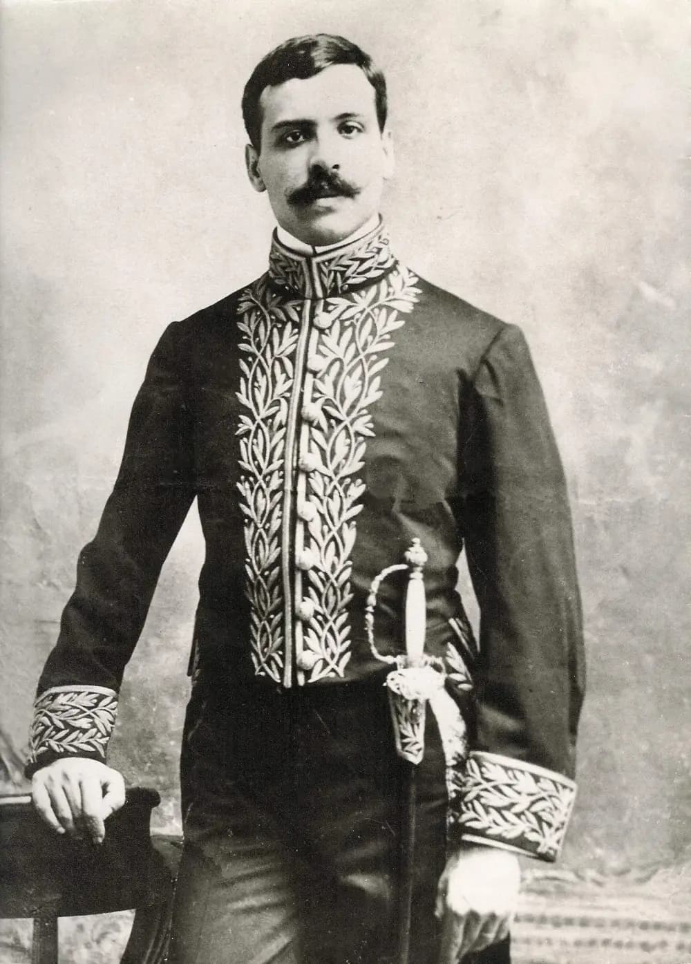 Black-and-white portrait of a young man with a dark moustache standing beside a chair, wearing a high-collared dark tunic embroidered with branches of leaves down the front and on the cuffs, with a sword at his side.