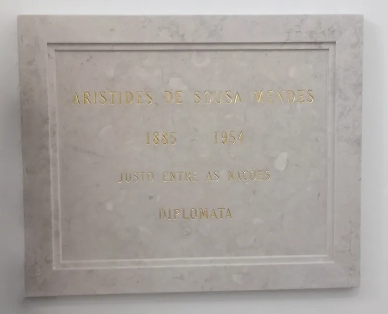 A rectangular pale stone plaque with a raised border, set into a white wall, with four lines of gilded capital letters: ARISTIDES DE SOUSA MENDES, 1885 – 1954, JUSTO ENTRE AS NAÇÕES, DIPLOMATA.