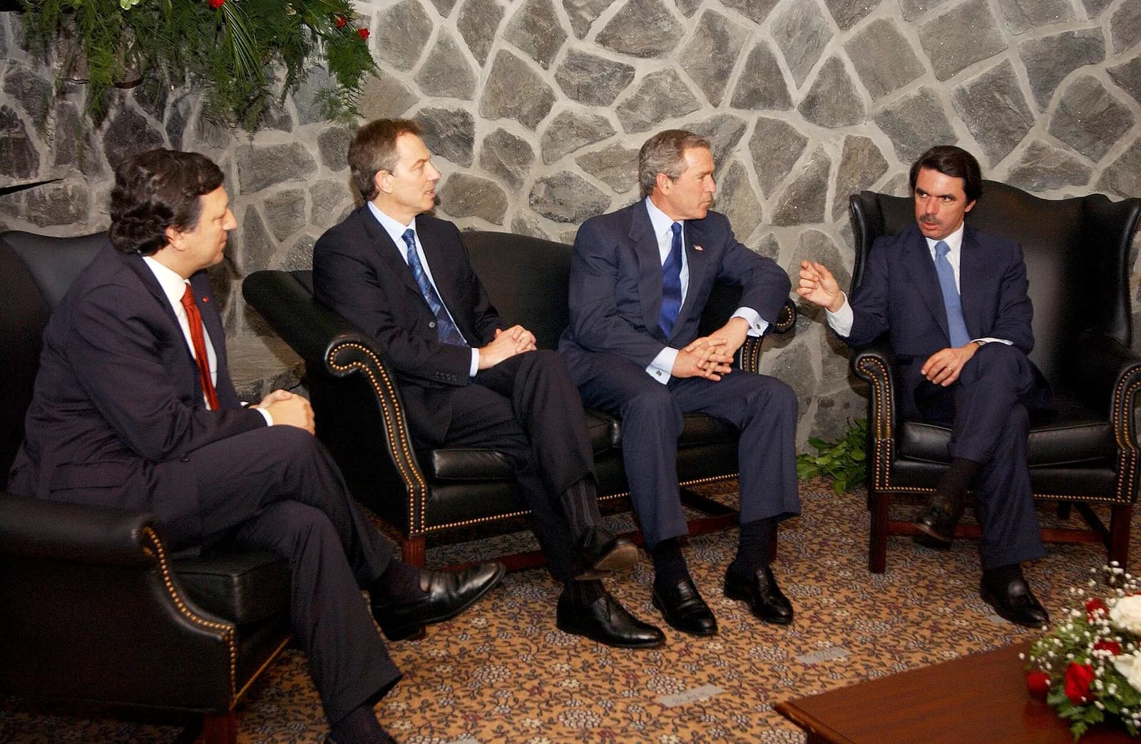 Four men in dark suits sit in black leather armchairs and a sofa in front of a stone wall; the man on the right gestures as he speaks while the others listen.