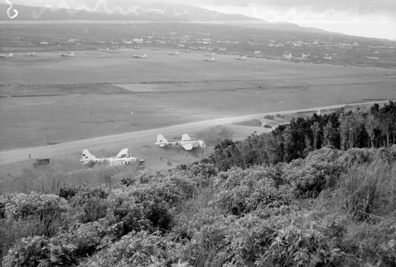 View from a scrubby hillside over a wide, flat airfield. Two large four-engined bombers are parked near a dirt track in the foreground and more aircraft stand in a line across the field, with scattered houses and hills beyond.