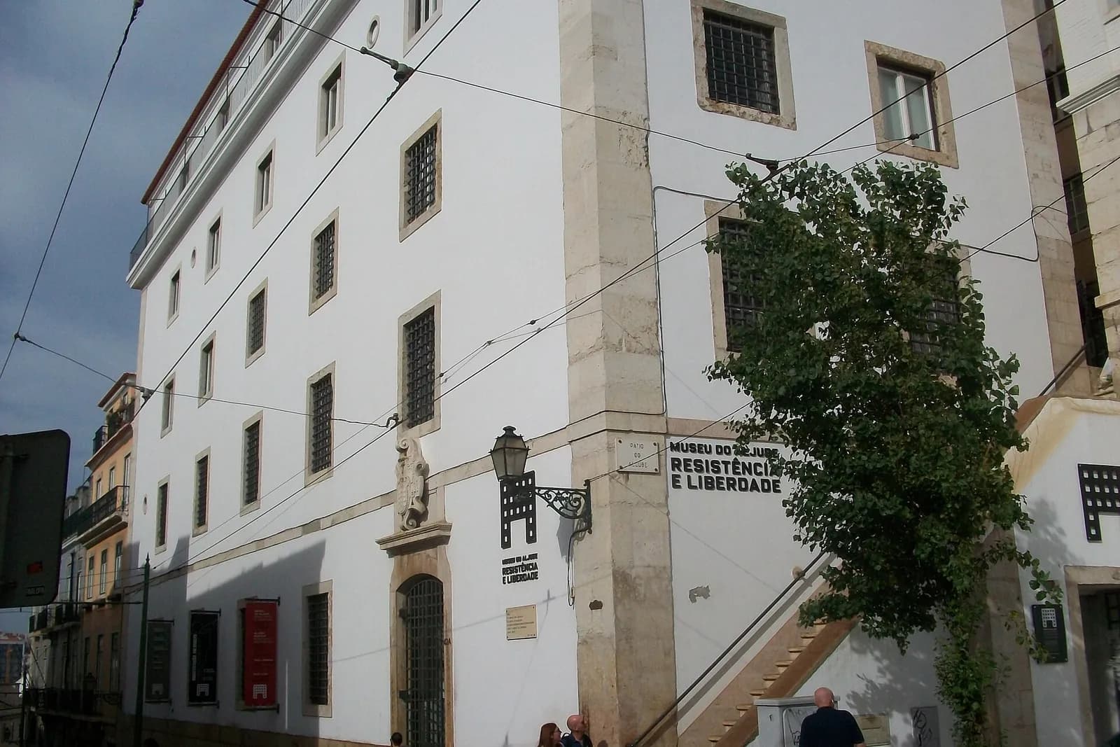 A tall white building with stone corners and small barred windows on a steep Lisbon street, with tram wires overhead and a sign on the wall reading Museu do Aljube Resistência e Liberdade.