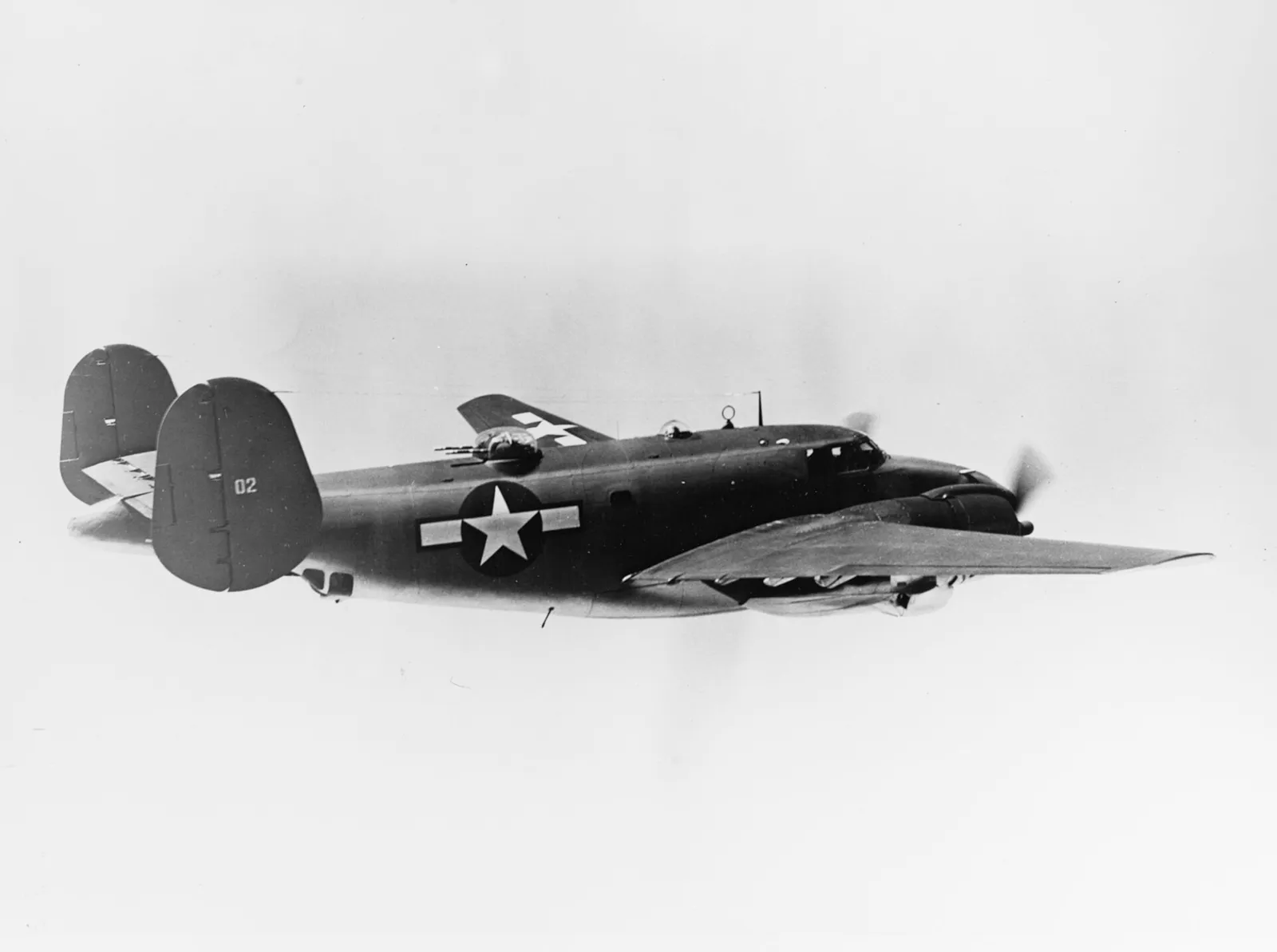 Black-and-white photograph of a dark twin-engined, twin-tailed aircraft with a gun turret on top of the fuselage and a white star insignia, flying against a pale sky.