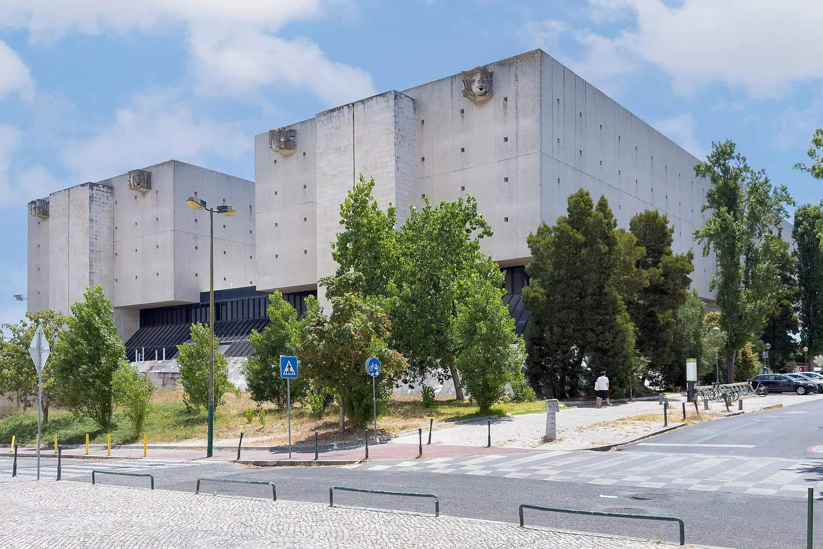 A massive pale concrete building made of blocky towers with small square openings and carved faces near the top, set behind trees beside a road and a pedestrian crossing under a blue sky.