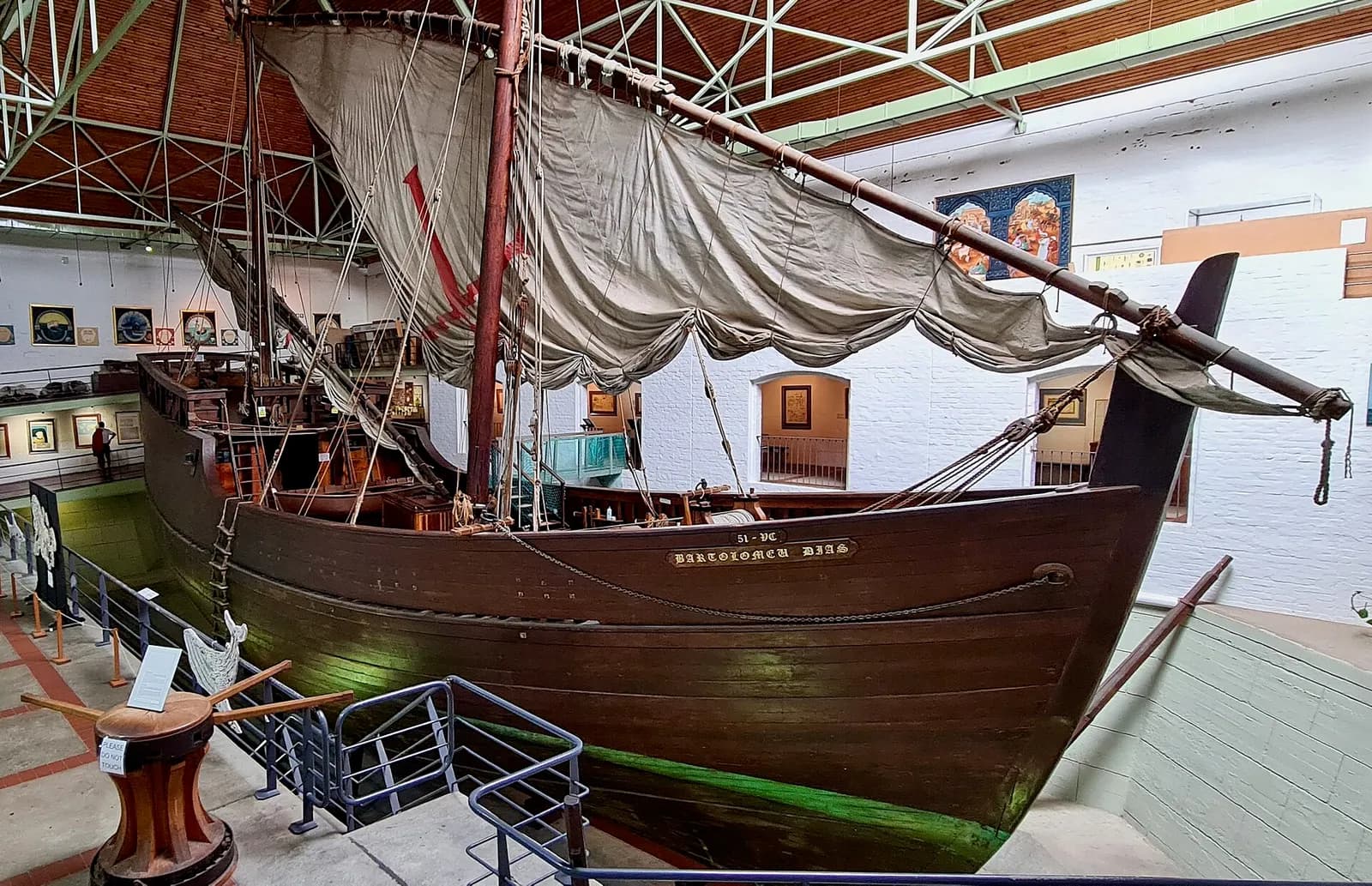 A wooden two-masted sailing ship with a long lateen yard and a furled sail painted with a red cross stands indoors in a dry pit in a large museum hall. The name Bartolomeu Dias is painted on a board on its bow.