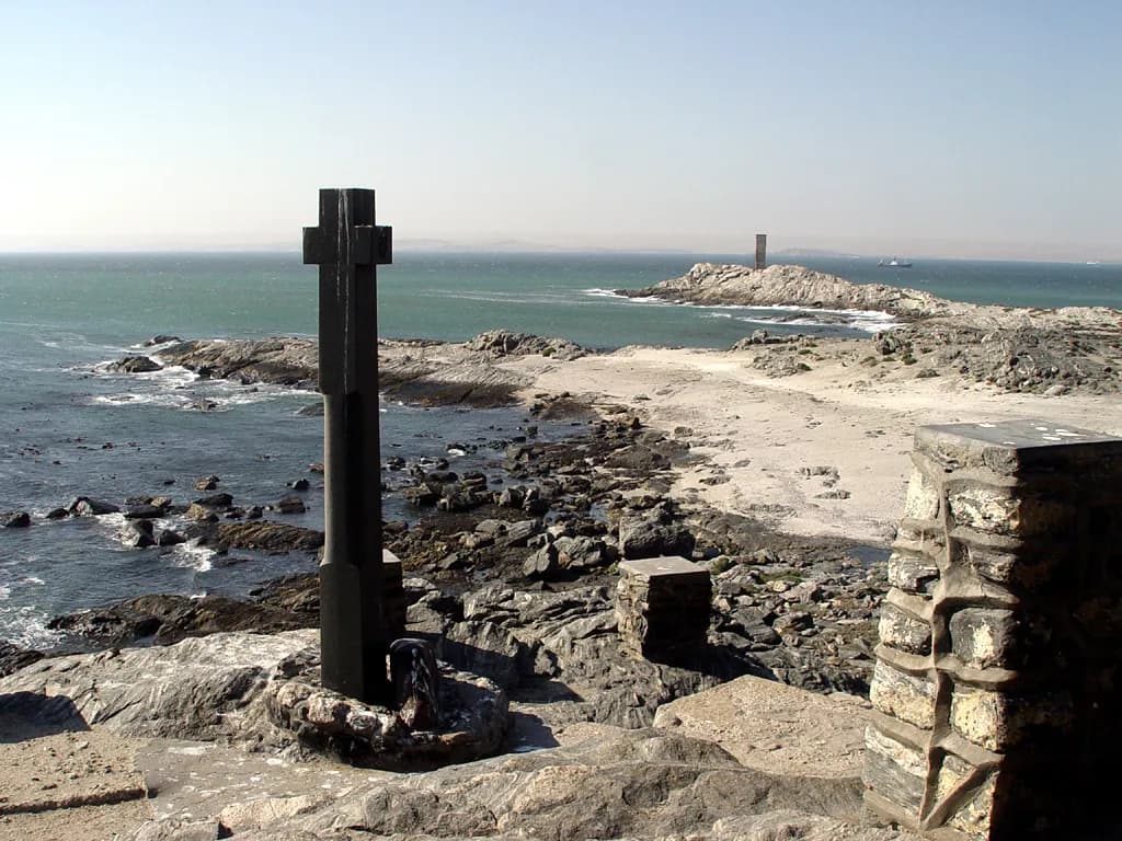 A tall dark stone cross on a rocky point above the sea, with a sandy cove and rocks behind it and a small stone tower on a further headland. A low stone pillar stands in the foreground on the right.