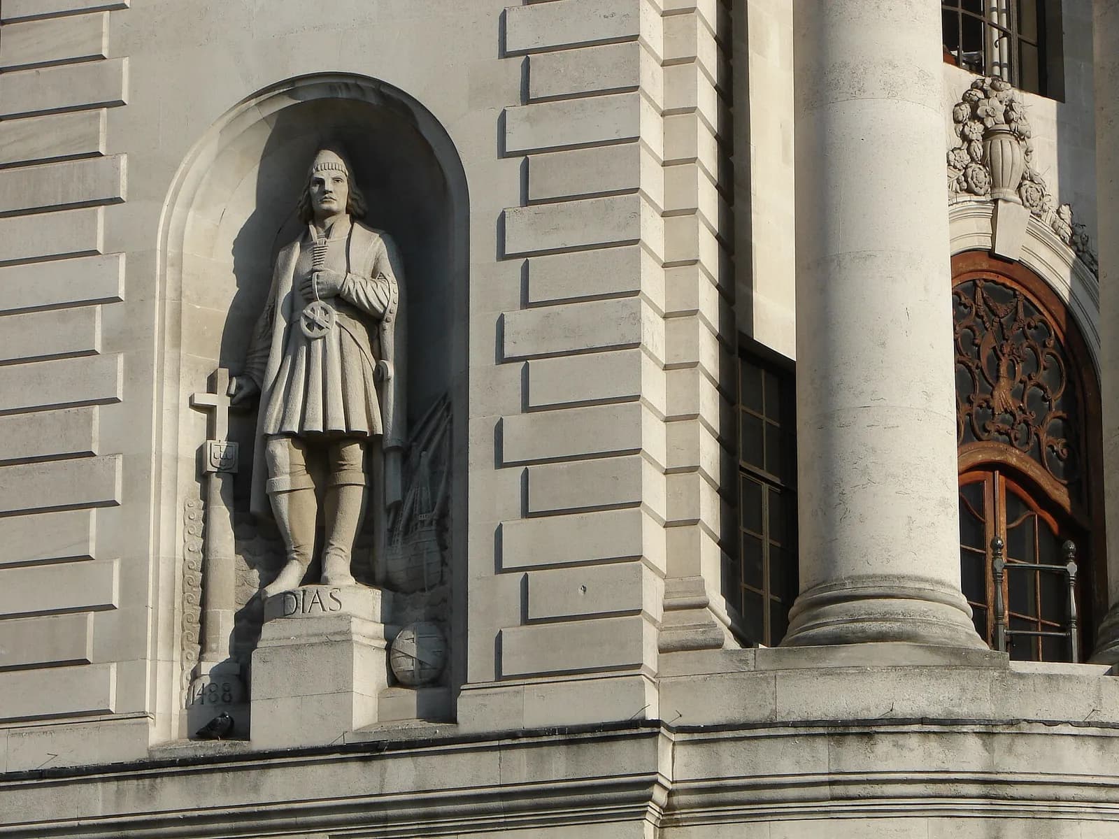 A stone statue of a man in a cap, tunic and boots stands in an arched niche in a pale stone facade. A cross-topped pillar with a coat of arms is carved beside him, the year 1488 on its base, and the name DIAS on his pedestal. A large column and an ornate wooden door are to the right.