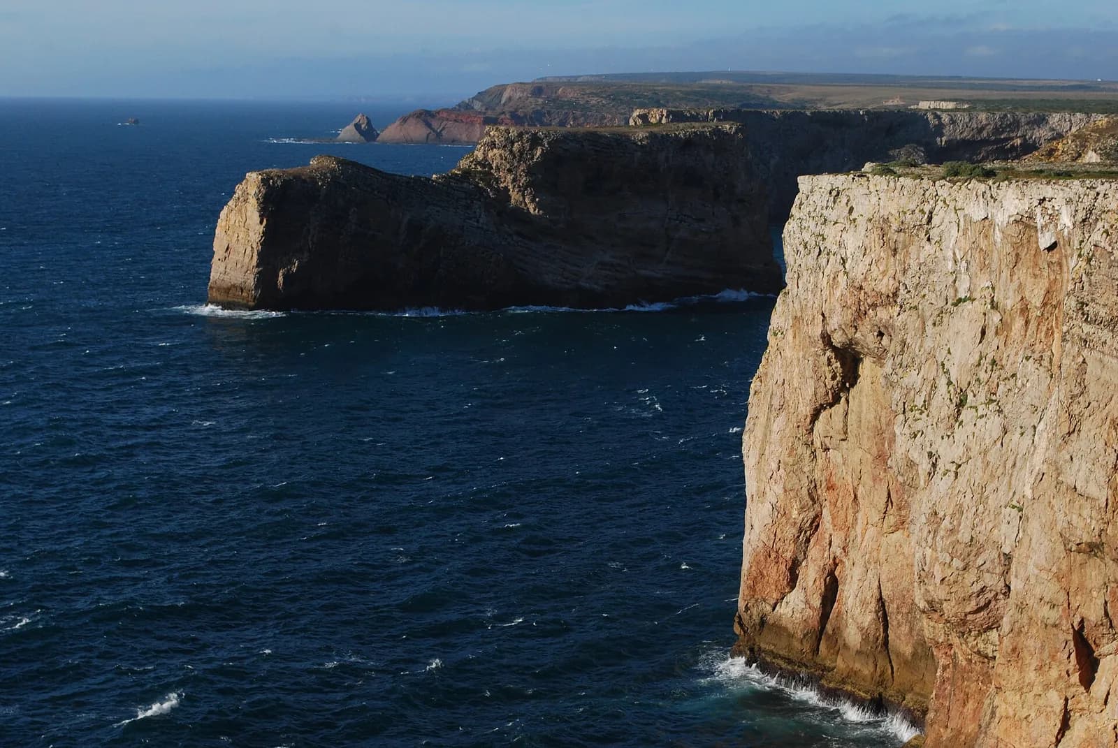 Sheer sandstone-coloured cliffs drop straight into a deep blue, wind-ruffled sea. Beyond them a flat-topped headland and a line of further cliffs stretch into the distance under a pale sky.