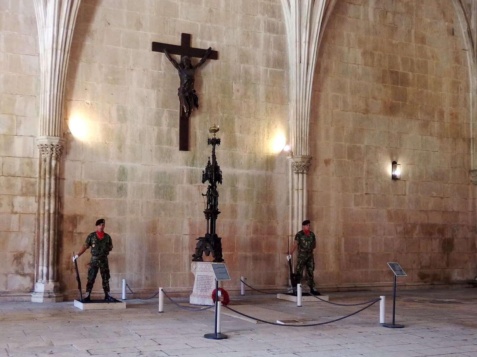 A vaulted stone hall with a dark, damaged bronze figure of Christ on a large wooden cross high on the wall. Below it a tall black ornamental lamp stands on a stone plinth with a red wreath, and two soldiers in camouflage, black berets and red neckerchiefs stand guard on either side behind a rope barrier.