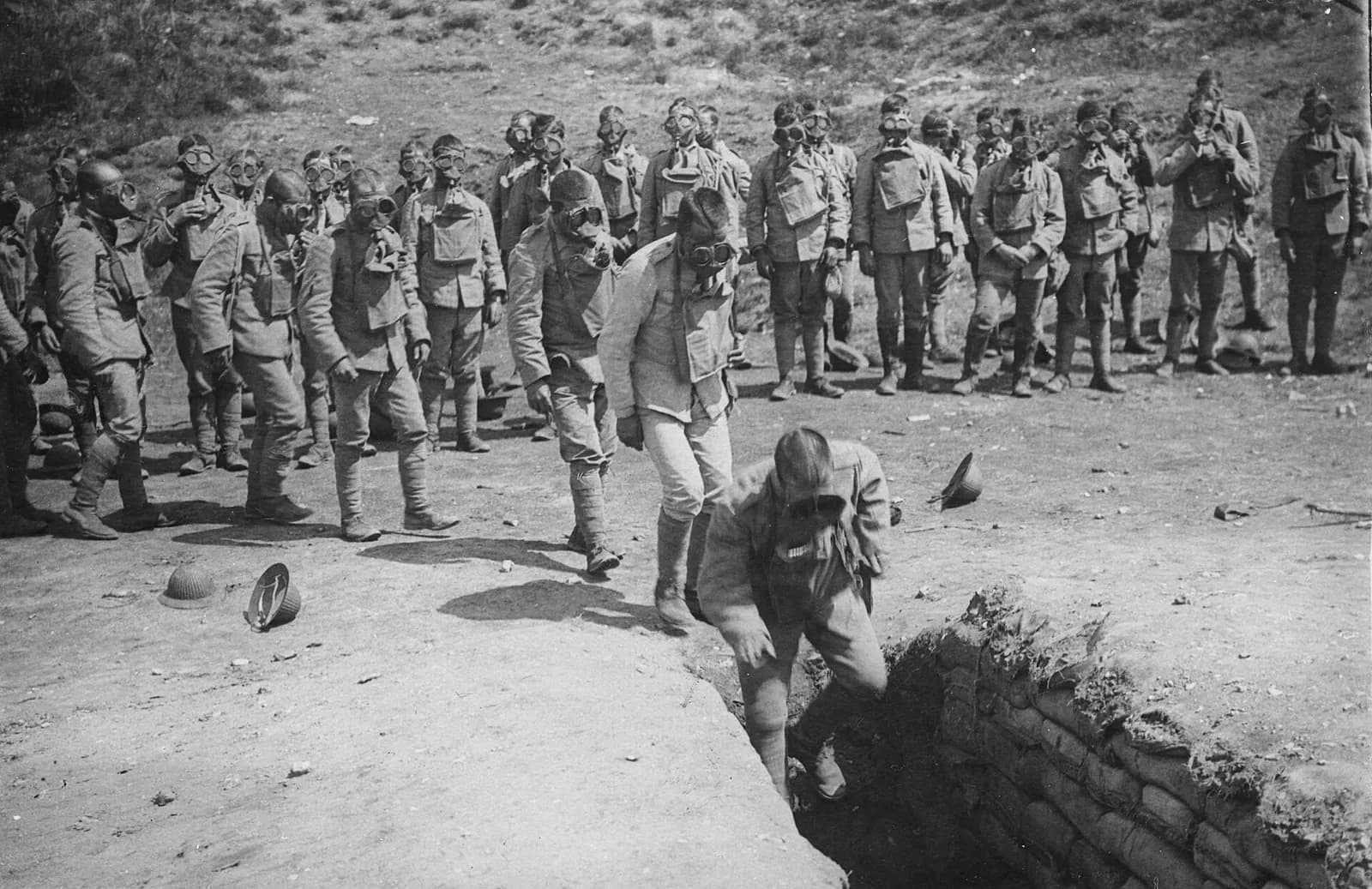 Soldiers in gas masks and pale tunics wait on bare, dusty ground while a file of them walks towards a narrow trench lined with sandbags, the first man climbing down into it. Two steel helmets lie on the ground in front of them.