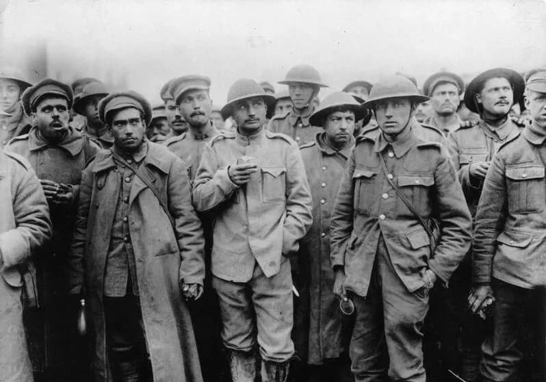 A crowd of captured soldiers stands packed together, staring towards the camera. One in front wears the Portuguese steel helmet with its ridged crown; others wear British-style flat-brimmed helmets or peaked caps, with long greatcoats or tunics. Their faces are tired and unshaven.
