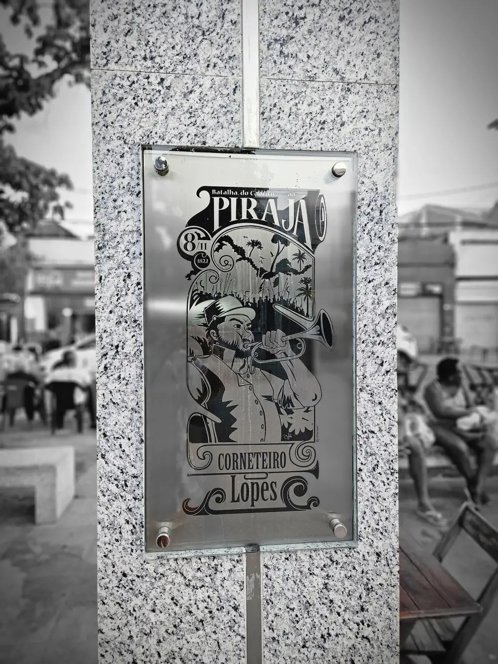 A metal plaque set into a granite pillar shows a drawing of a bearded man in a cap blowing a bugle in front of palm trees and a crowd, with the words Batalha do Cabrito ao Pirajá, 8/11/1822, Corneteiro Lopes.