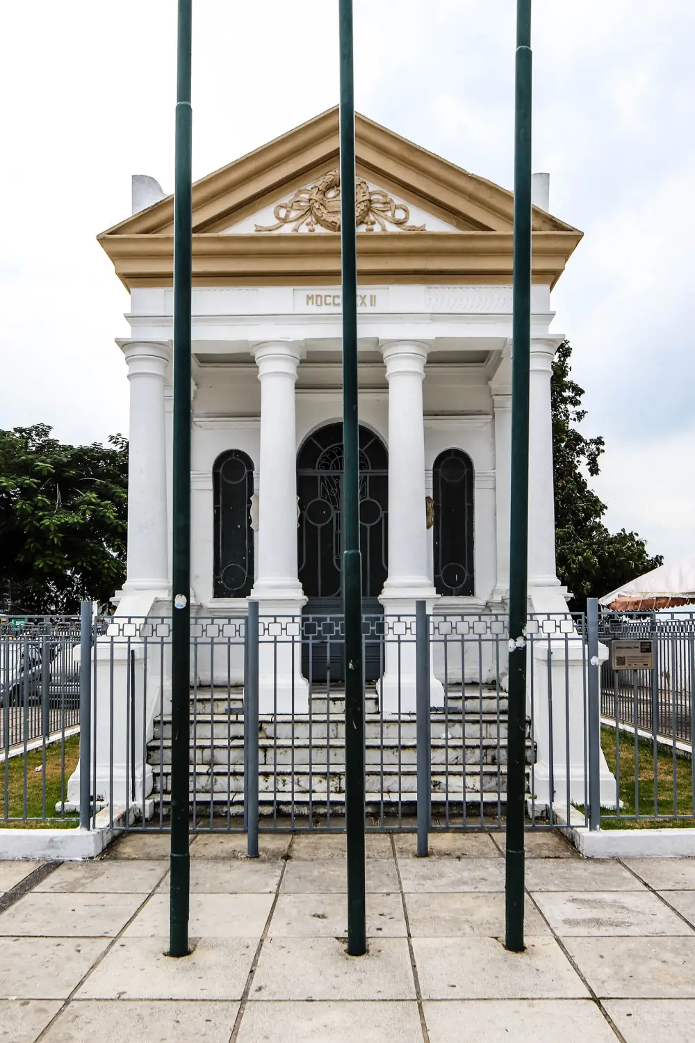 A small white neoclassical temple with four columns, a triangular pediment carved with a wreath and the date MDCCCXXII, behind railings and three tall flagpoles.