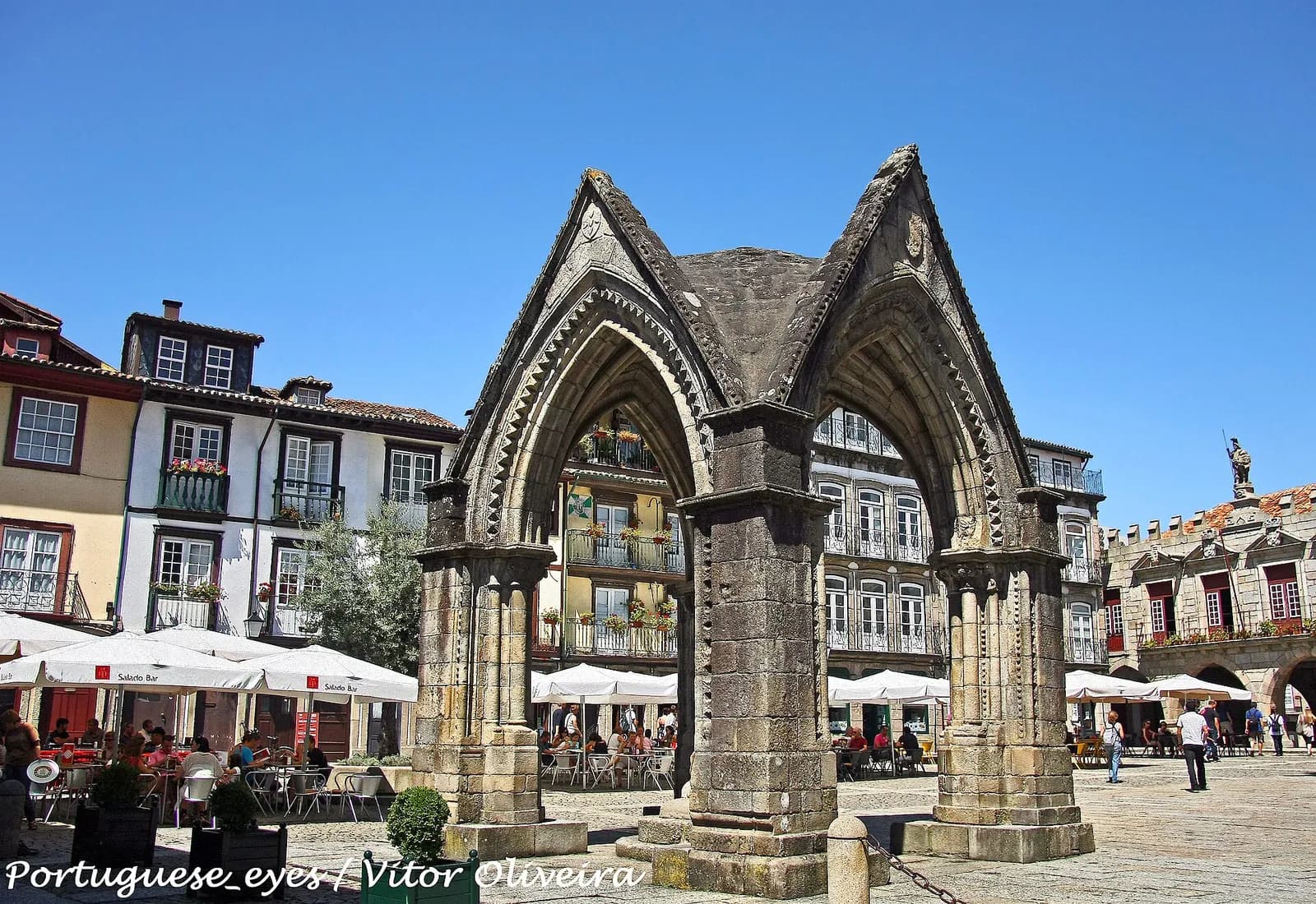 A weathered granite Gothic structure in a sunny town square: pointed arches with zigzag mouldings on stout piers with attached shafts, topped by steep triangular gables, with café umbrellas and old houses with flower-filled balconies behind.