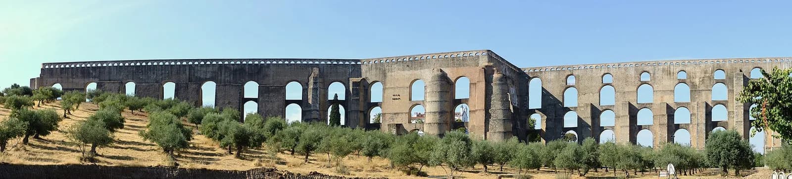 A panorama of a long stone aqueduct on tall arches, in places stacked two and three tiers high, rising behind rows of olive trees on dry grass under a blue sky.