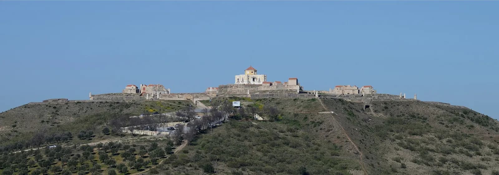 A panorama of a hilltop fortress: long low stone walls run along the crest of a scrubby hill planted with olive trees, with a pale house rising in the middle, under a clear blue sky.