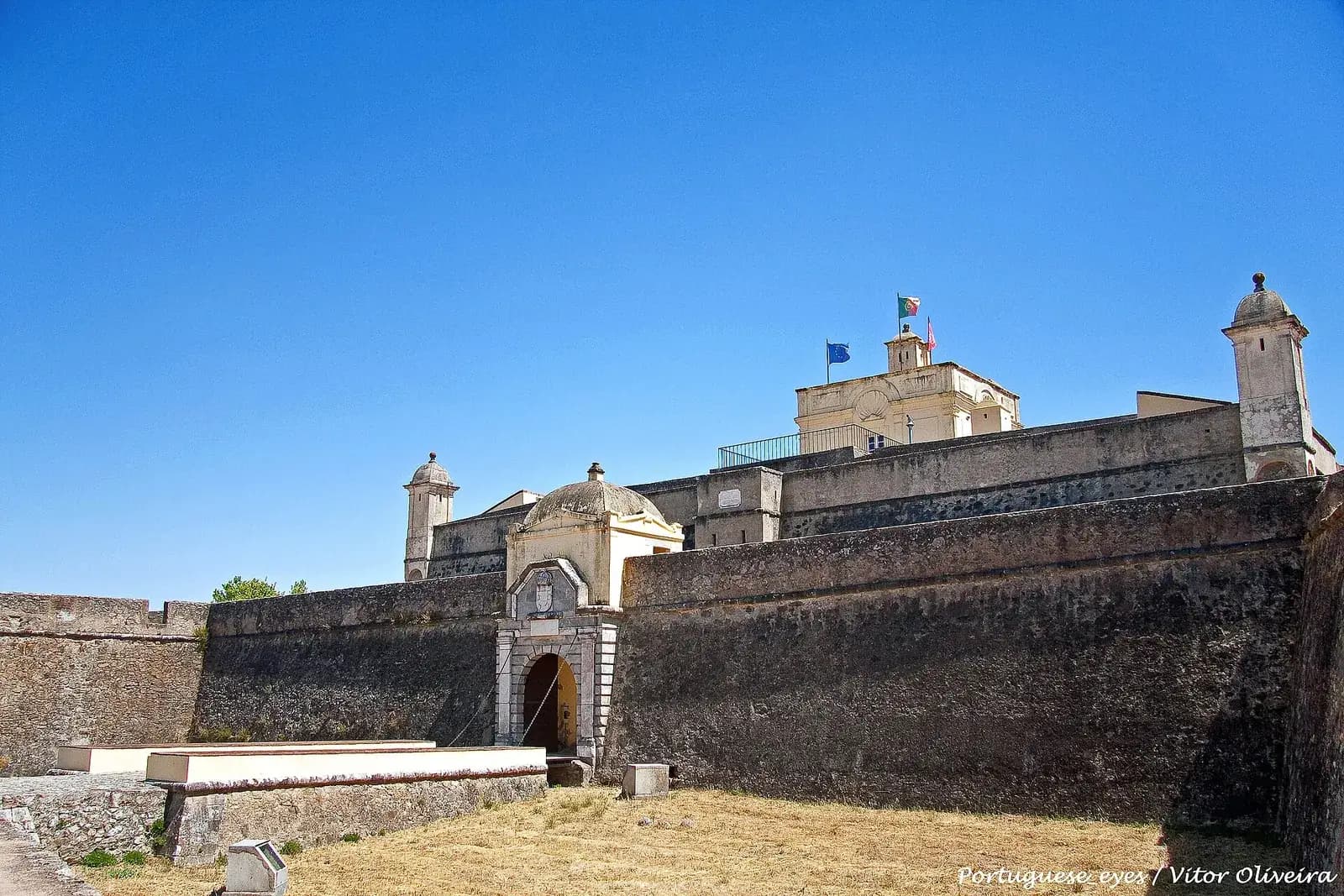 The gate of a stone fortress seen from its dry ditch: a carved stone doorway with a small domed roof in a long, sloping grey wall, with corner watchtowers and a pale building flying the Portuguese and European flags above.