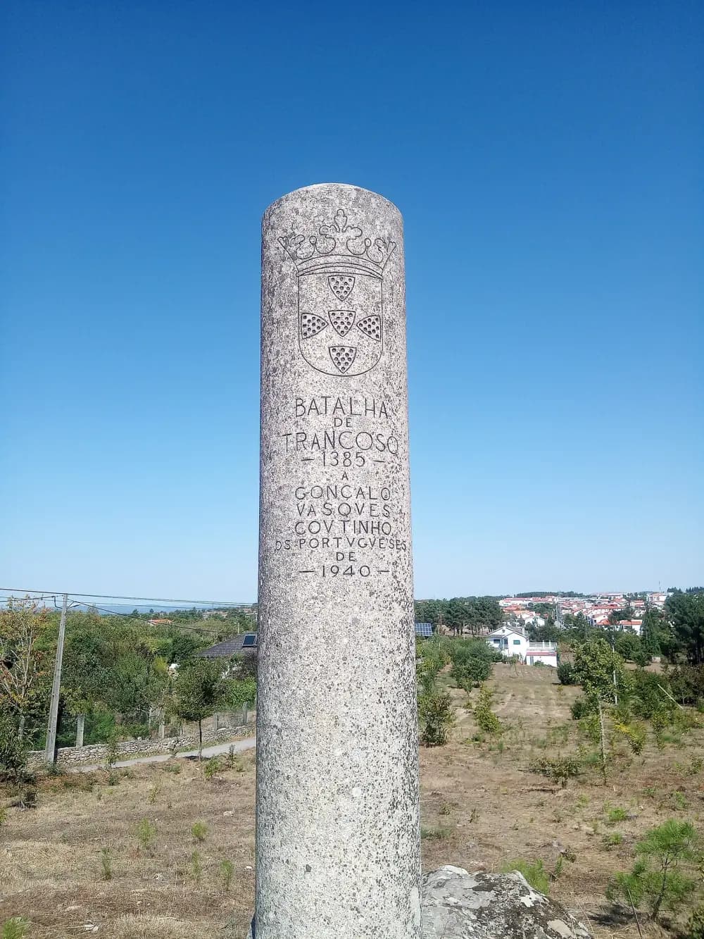 A tall granite column carved with the royal arms of Portugal and the inscription Batalha de Trancoso 1385, a Gonçalo Vasques Coutinho, os Portugueses de 1940, standing in scrubland with the town of Trancoso in the distance.