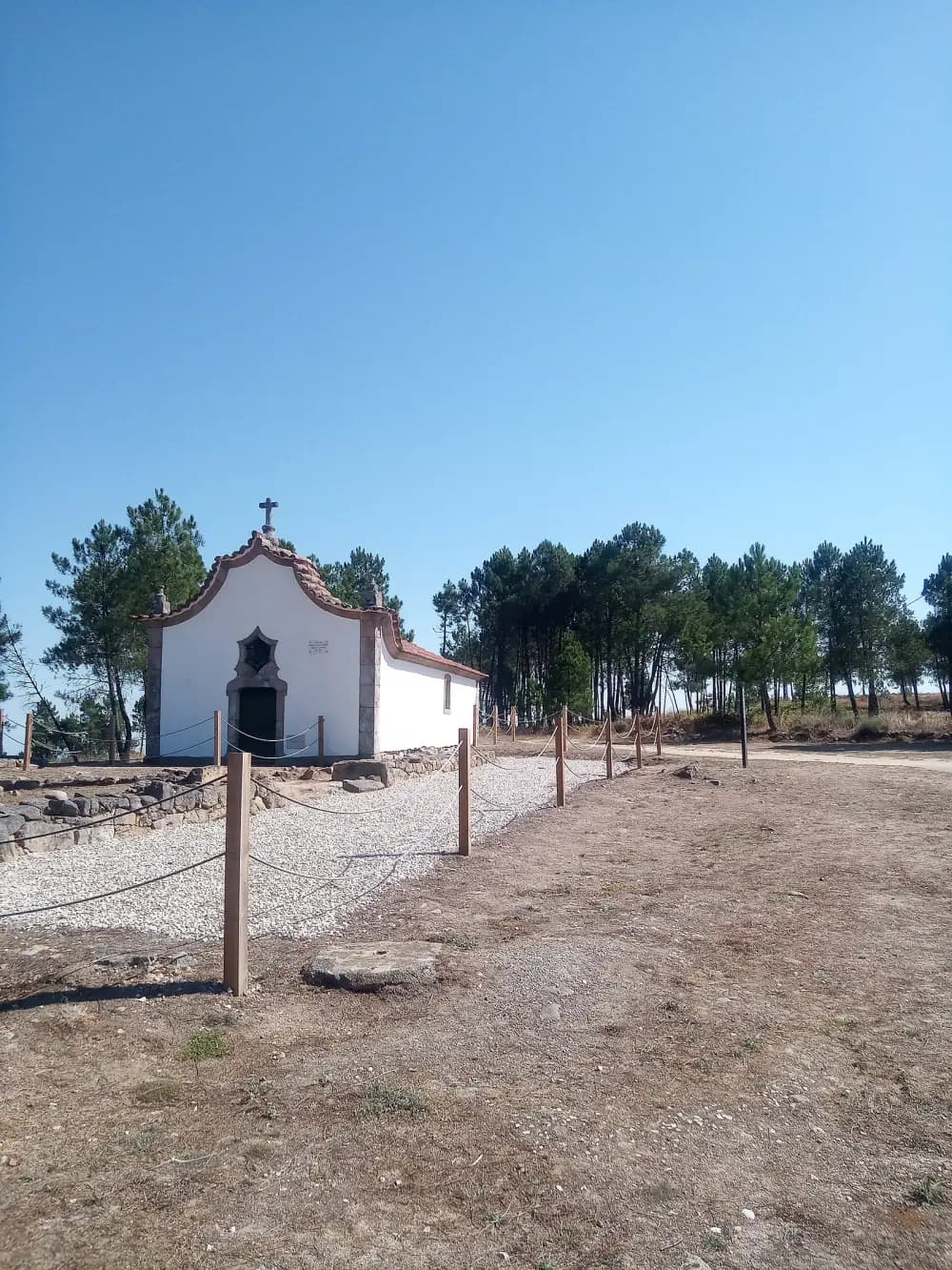 A small whitewashed chapel with a curved granite gable and a stone cross stands among pine trees. In the foreground, low stone foundations inside a gravel area are fenced off with wooden posts and rope.