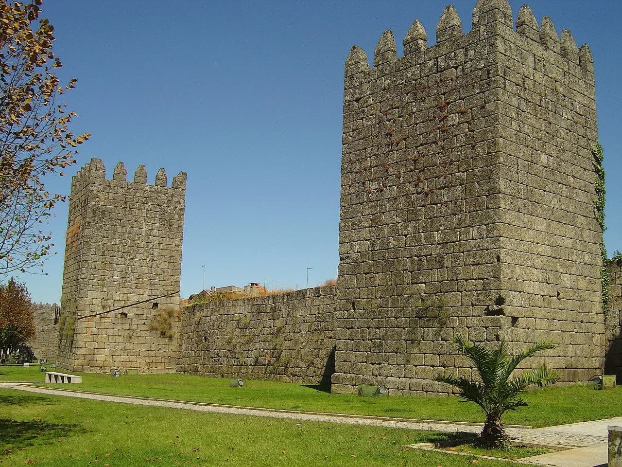 Two tall square granite towers with pointed merlons, joined by a stretch of curtain wall, stand on a lawn under a clear blue sky.