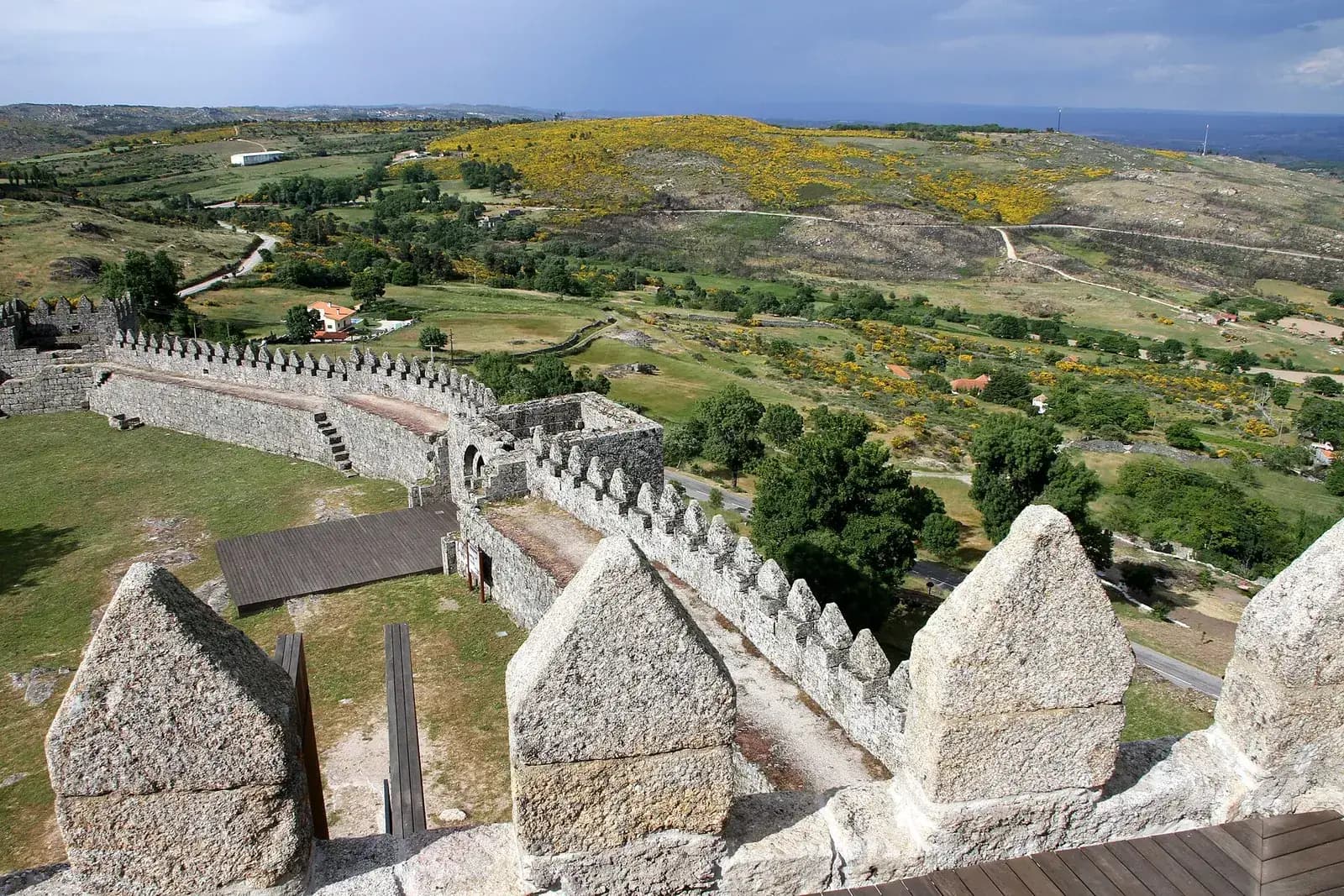 Crenellated granite walls run along a slope below a stone parapet, with fields, scattered trees, a road and hills covered in yellow broom stretching away under a grey sky.