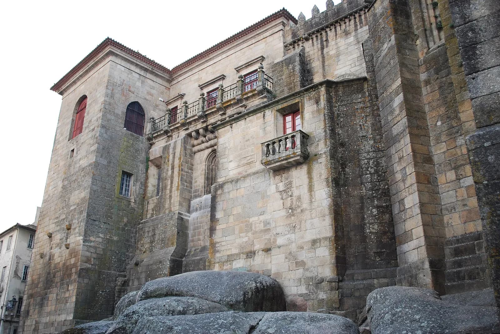 The rear of a large granite cathedral rises above rounded boulders, with a tall square tower on the left, crenellations along the roofline and later windows and balconies set into the old walls.
