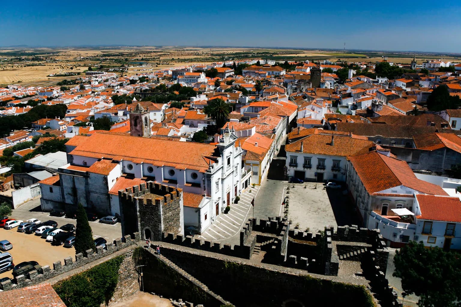 Aerial view over the whitewashed houses and orange-tiled roofs of Beja, with the battlemented walls of the castle in the foreground and the flat, dry plain of the Alentejo stretching to the horizon.
