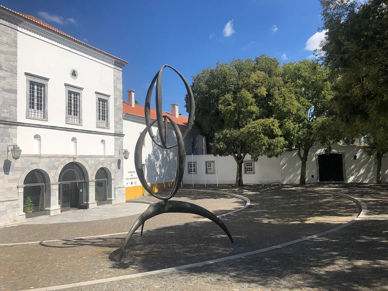 A tall, dark bronze sculpture of interlocking loops on curved legs stands in a cobbled square, in front of a white and grey stone building with arched doorways and a row of trees.