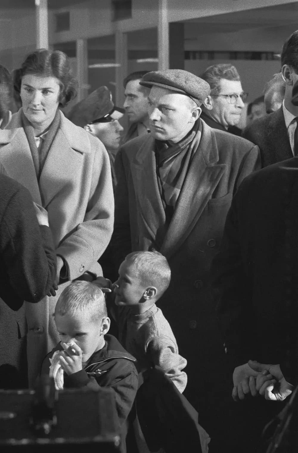 Black-and-white photograph of a crowd of travellers in winter coats in an airport hall: a woman in a light coat, a young man in a flat cap, and two small boys in front of them, one holding a handkerchief to his face.