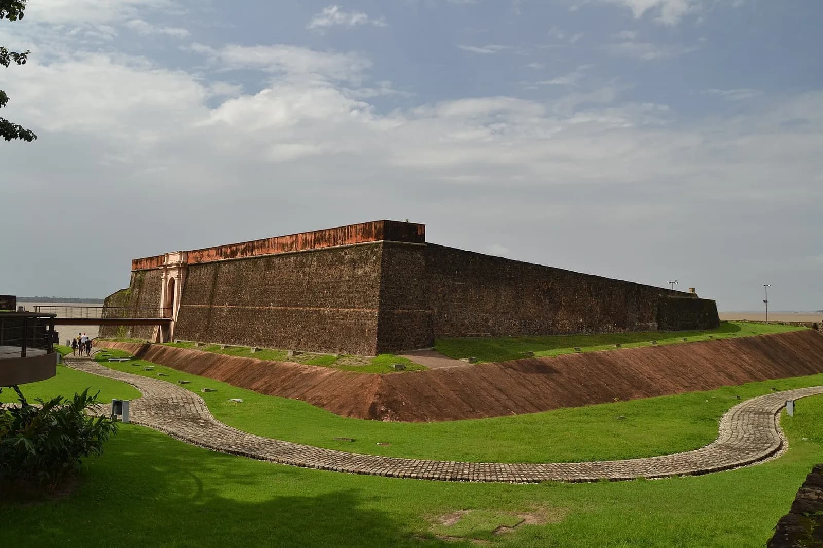 Low, dark stone walls of a fort rise from a lawn beside a wide brown river under a cloudy sky, with a curving path leading towards them.