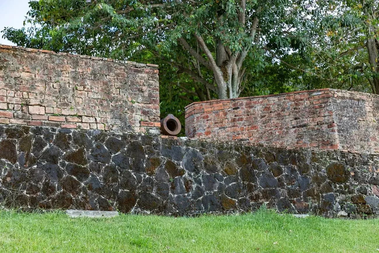 A rampart of dark, irregular stones set in pale mortar, topped by a parapet of weathered red brick. The muzzle of an old cannon pokes through a gap in the parapet, with a large tree behind.