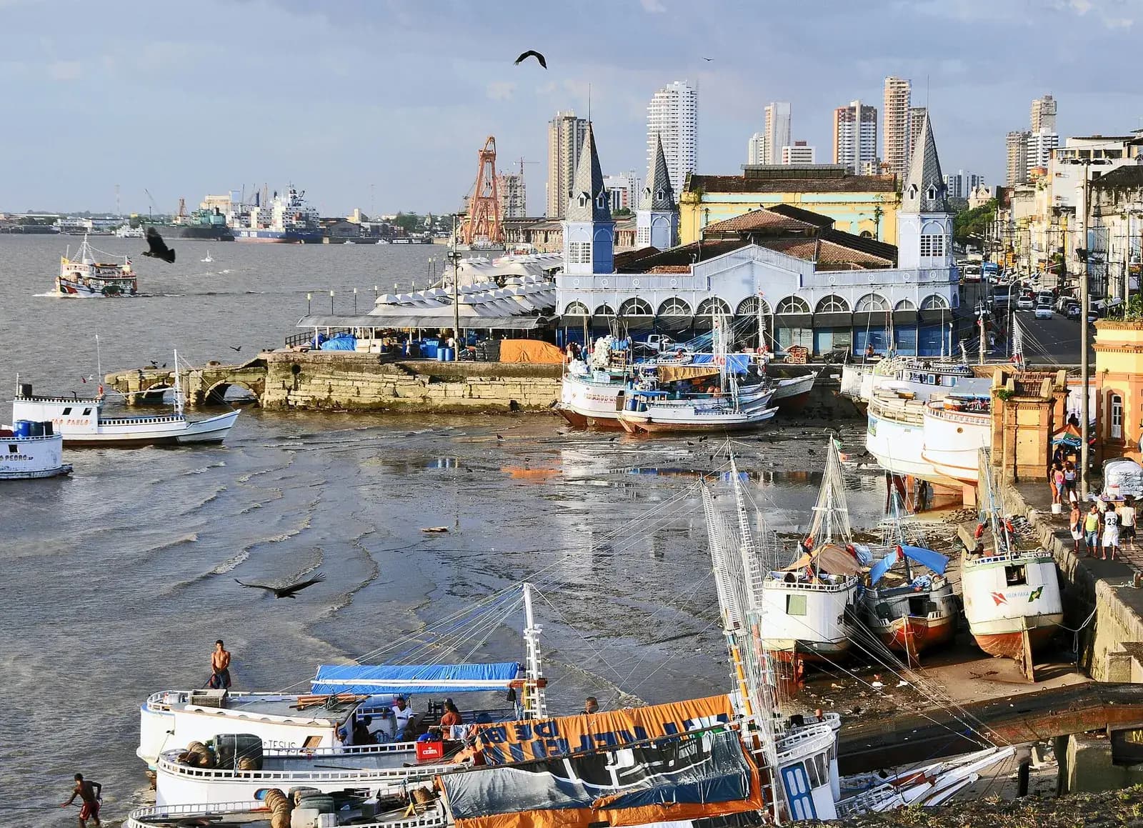 Fishing boats lie on the mud of a harbour at low tide in front of a long pale-blue market hall with pointed corner turrets; high-rise towers and port cranes stand behind it under a cloudy sky.