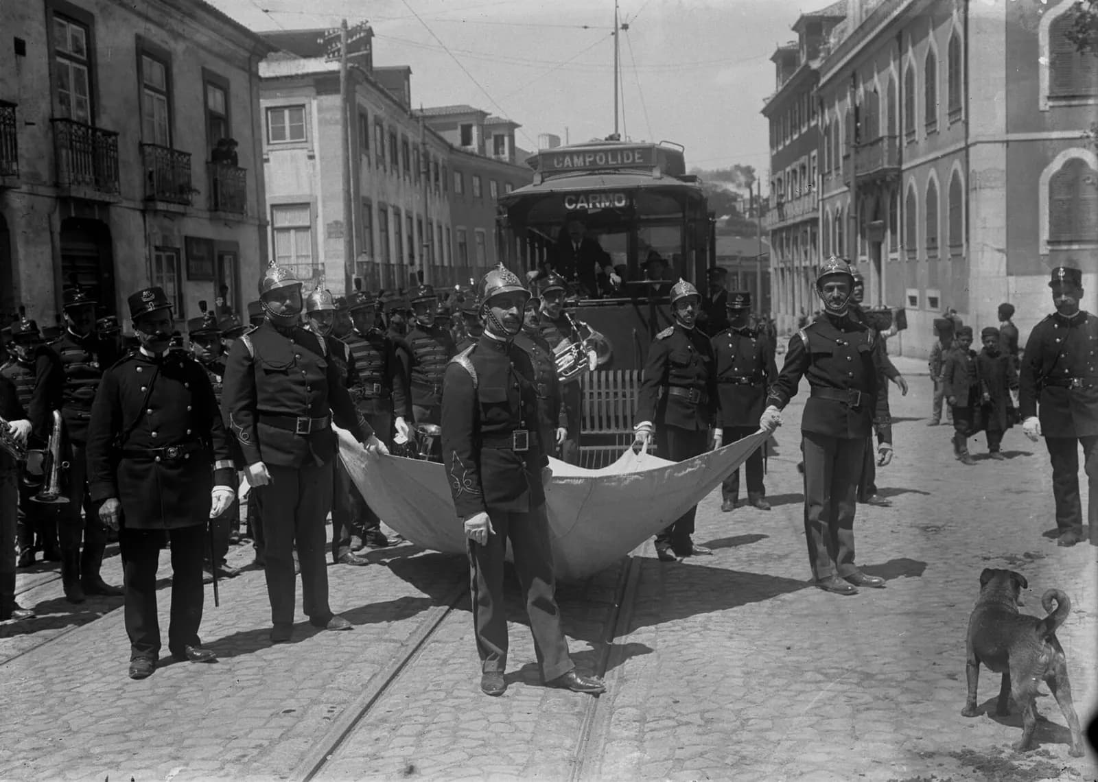 Firemen in brass helmets and dark uniforms hold a large pale sheet stretched across a cobbled street with tram rails. Behind them stand bandsmen with brass instruments and a tram showing the destinations Campolide and Carmo; a dog watches in the foreground.