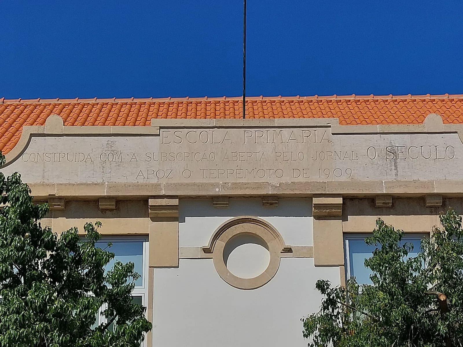 The top of a pale building with an orange tiled roof and a stone frieze carved in capital letters, above a round window, framed by two trees against a deep blue sky.