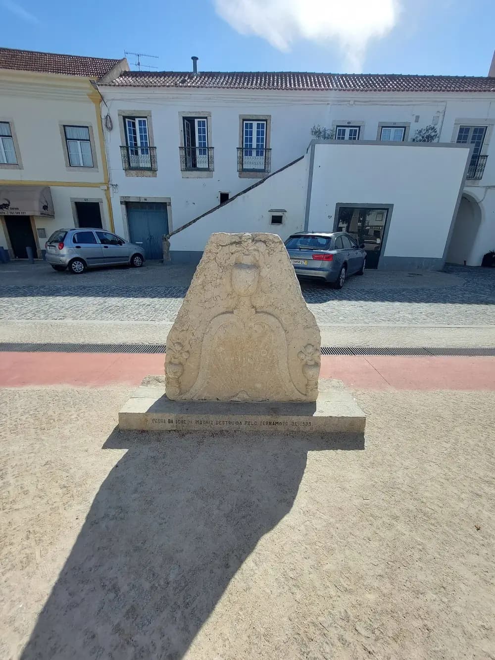 A carved limestone fragment with a shield-shaped panel and scrolled sides stands on a low plinth in a sandy square, in front of a row of white two-storey houses with parked cars.