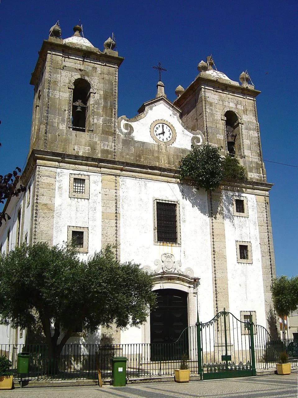 A white church façade with two stone bell towers, a clock in the central gable and a wrought-iron gate in front, under a deep blue sky.