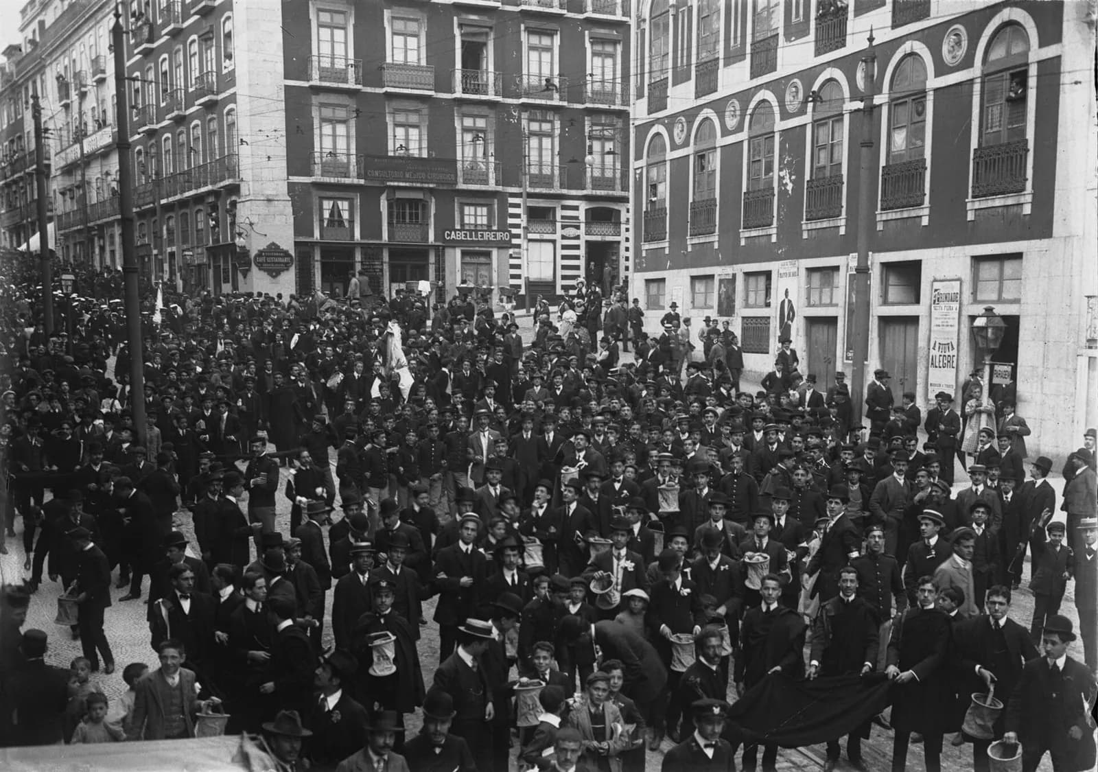 A dense crowd of young men in dark suits, boaters and bowler hats fills a street between tall buildings, where a poster advertises the operetta A Viúva Alegre. In front, many hold up collecting bags and a few stretch a dark cloth between them.
