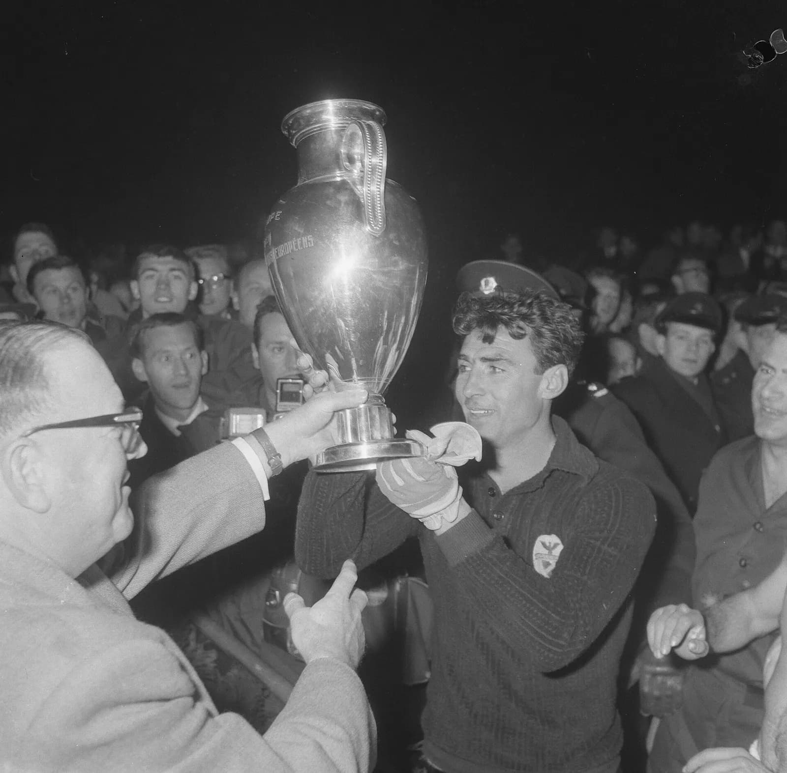 A curly-haired man in a dark goalkeeper's jersey with a club crest and white gloves grips a tall silver trophy with large handles, which an older man in glasses also holds. A crowd of young men and a policeman in a peaked cap look on.