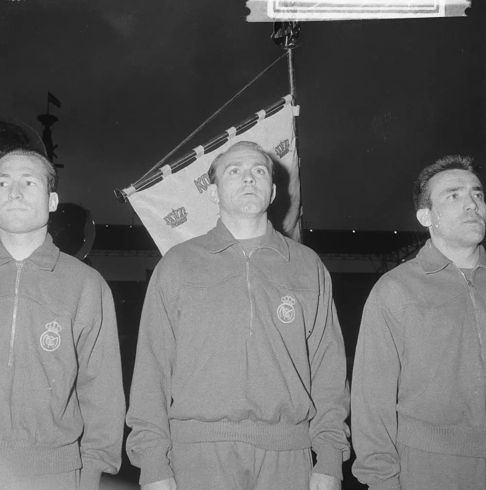 Three footballers in zipped tracksuit tops with a crowned club badge stand in a line looking up, in front of a hanging pennant with crowns on it. The man in the centre is balding.