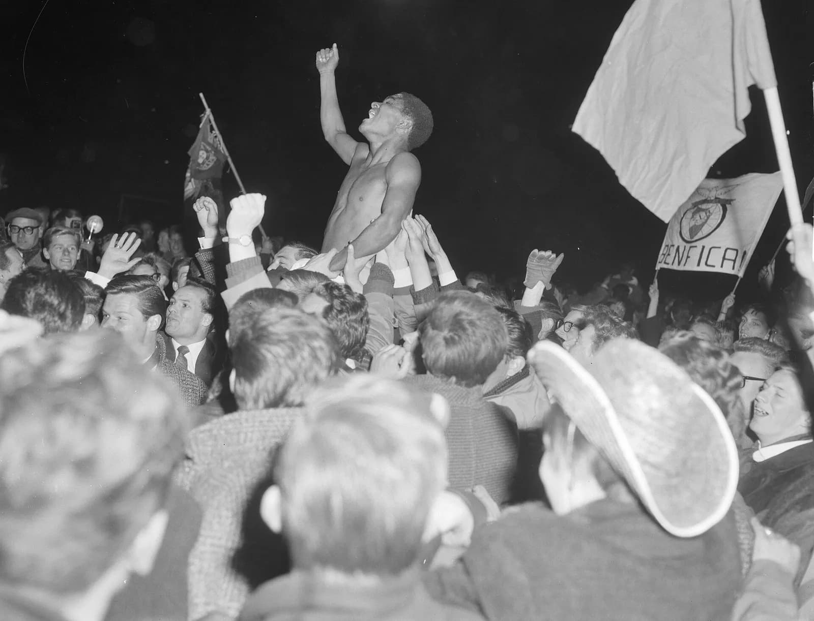 At night, a young Black footballer stripped to the waist is held up above a dense crowd of cheering young men, one fist raised and his head thrown back as he shouts. Behind him supporters wave flags, one a banner with a crest and the word Benfica.