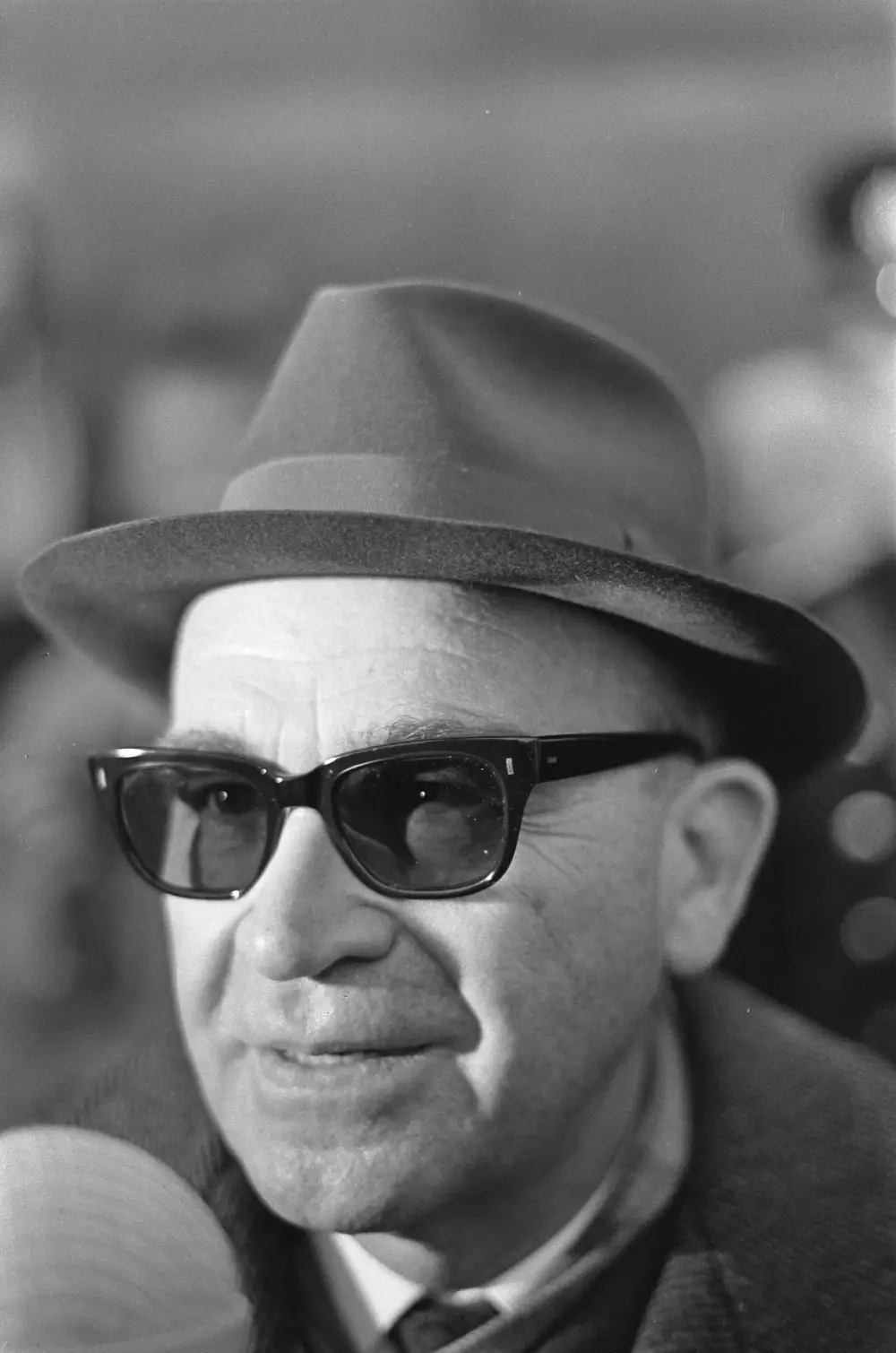 Close portrait of an older man in a felt hat with a dark band and thick-rimmed dark glasses, speaking, with a pale microphone at the lower edge of the picture.