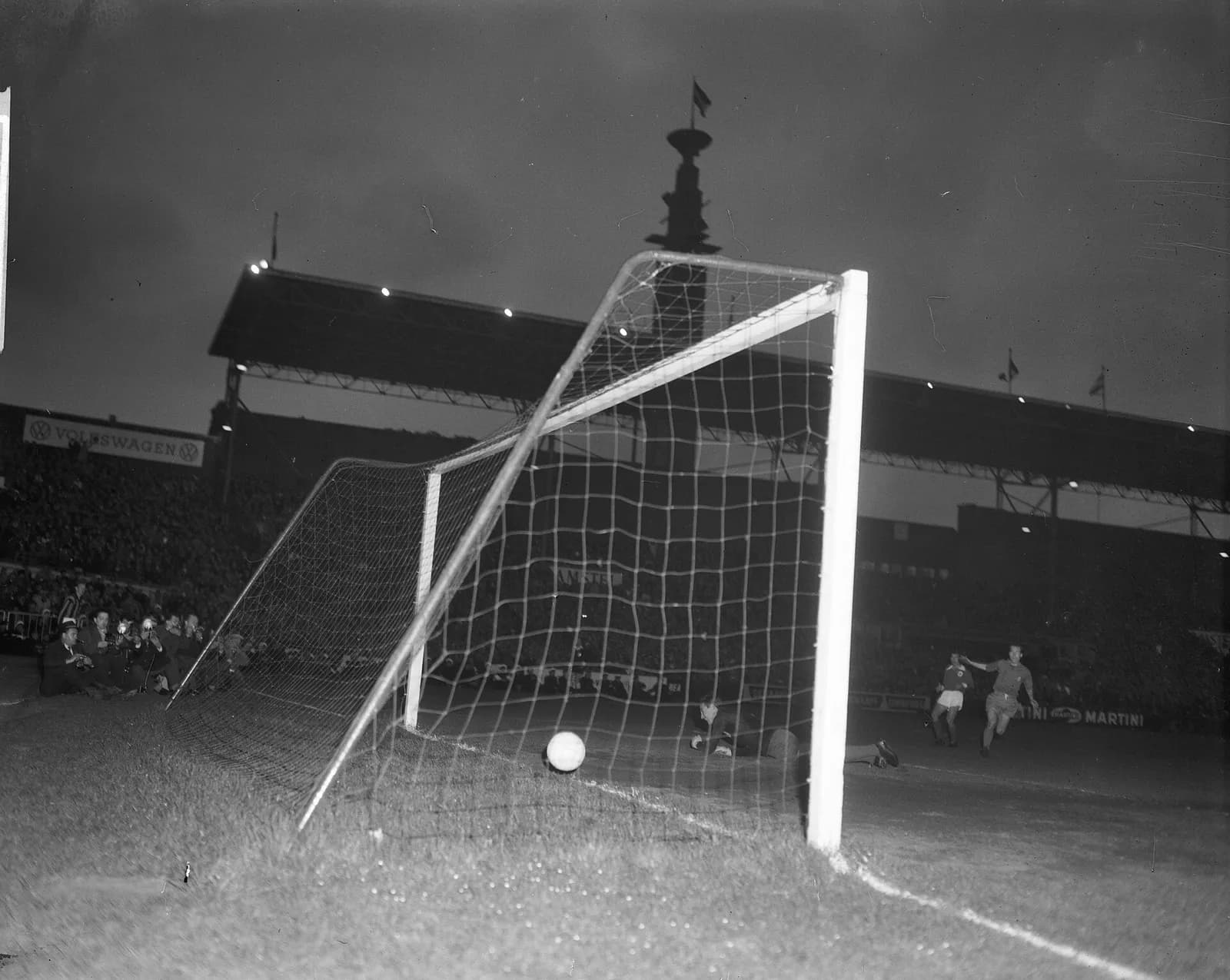 Floodlit night photograph taken from beside a goalpost: a football lies in the back of the net, a goalkeeper is on the ground near the goal line and two players run off in the distance, one with an arm outstretched. Photographers crouch by the net, and a covered stand and a tall tower rise behind.