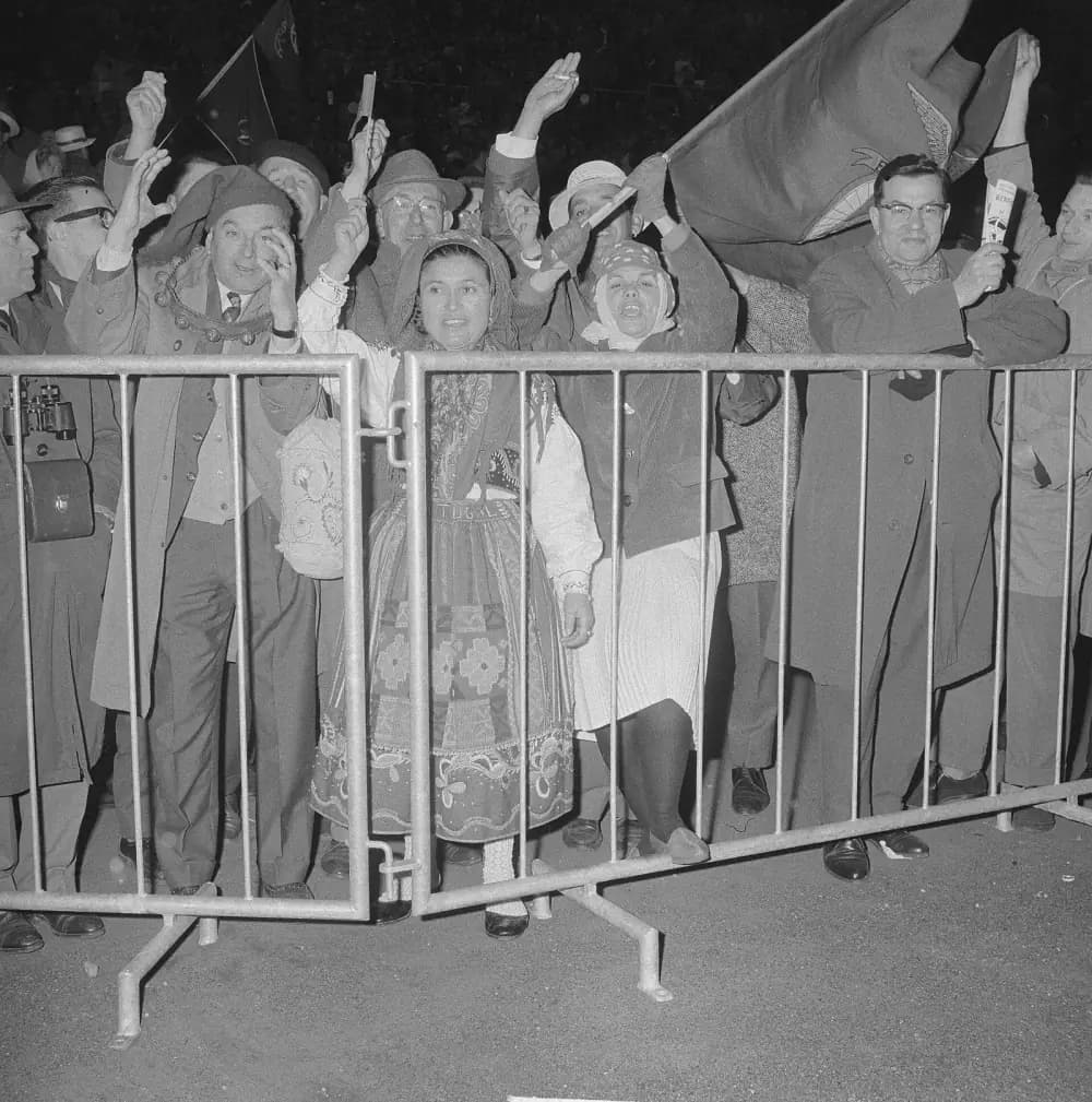 Supporters press against metal crowd barriers at night, waving and shouting. In front, a woman in a headscarf wears a traditional embroidered skirt and apron with the word Portugal stitched on it. A man beside her wears a long stocking cap, and a large flag with a club crest flies above them.