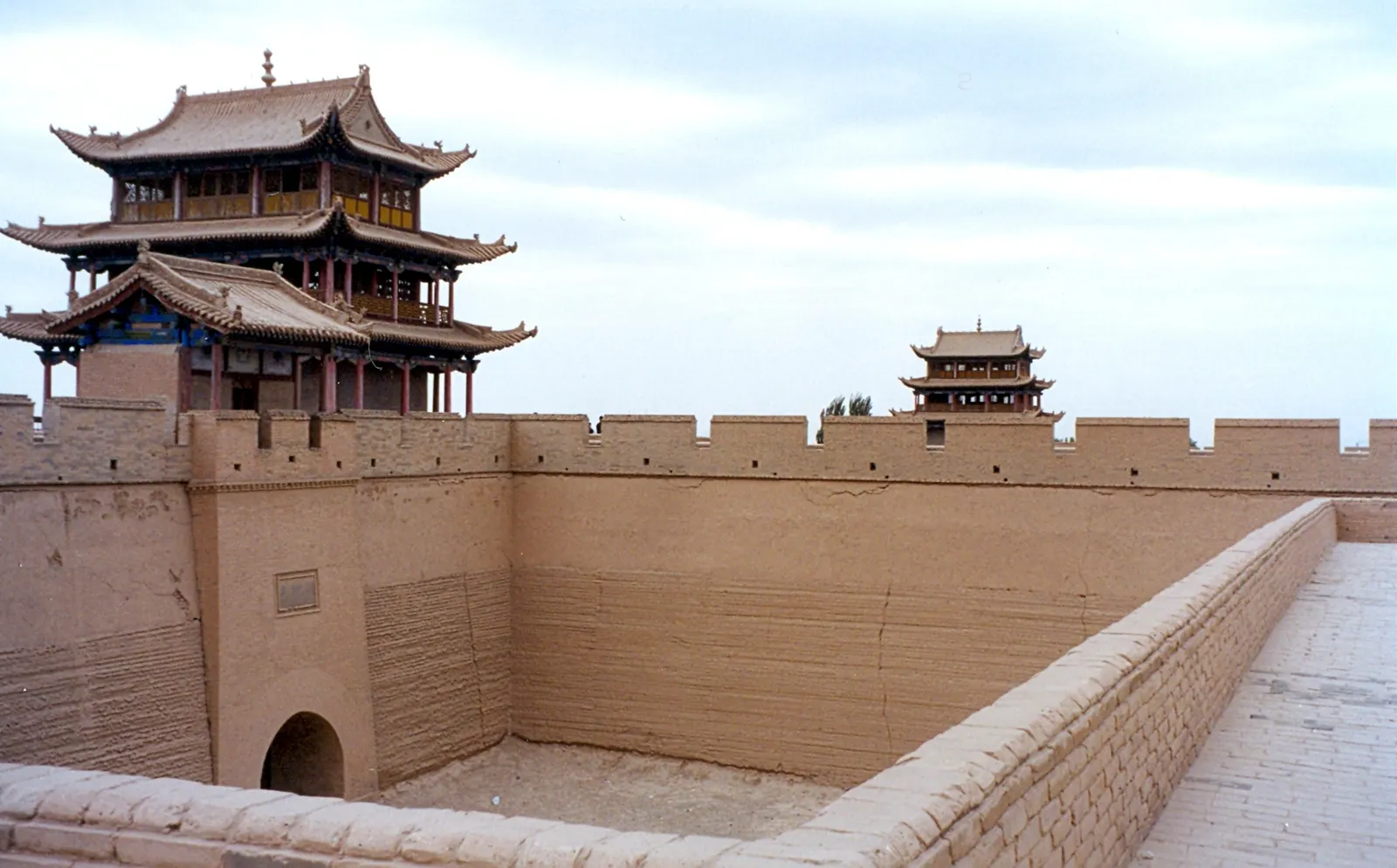 Thick rammed-earth walls with crenellated tops enclose an empty courtyard with an arched gateway. A three-storey Chinese gate tower with curved tiled roofs rises on the left, and a smaller tower stands on the wall in the distance.