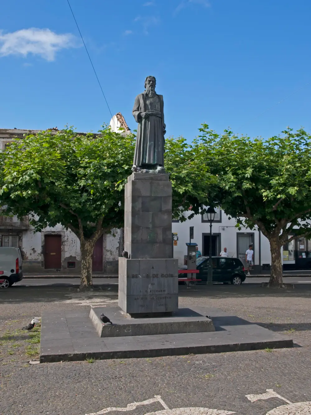 A bronze statue of a bearded man in a long belted robe stands on a tall dark stone pedestal in a cobbled square, framed by plane trees and low white houses. The inscription on the base reads Bento de Góis, 1562 to 1962.