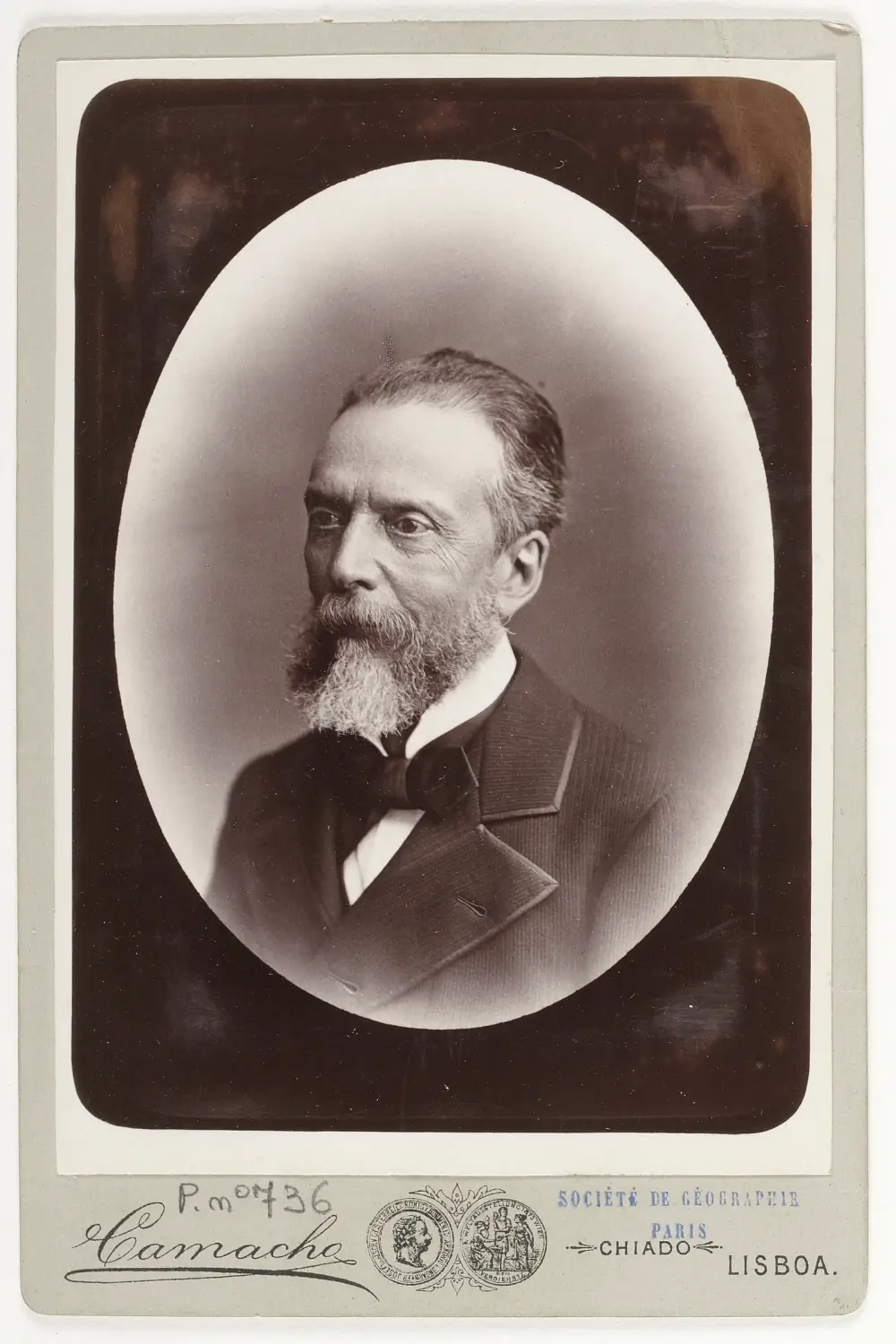 Studio portrait in an oval frame of an older man with swept-back grey hair and a full grey beard, wearing a dark jacket and a wide black bow tie, on a card mount printed with the photographer's name and Lisbon.