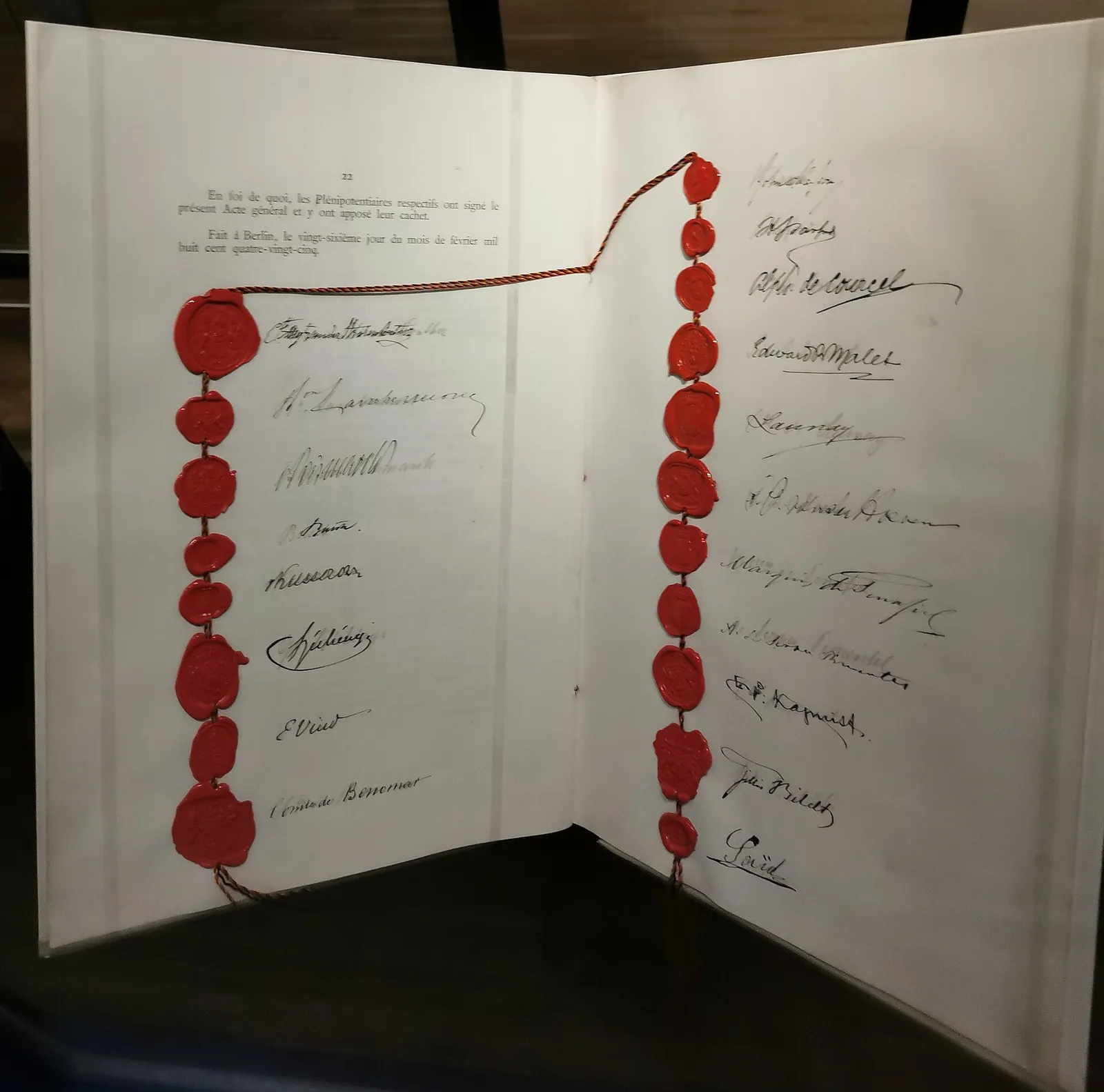 An open bound document in a display case. A printed French closing formula on the left page is followed by two columns of handwritten signatures, each beside a red wax seal, joined by a cord running down both pages.